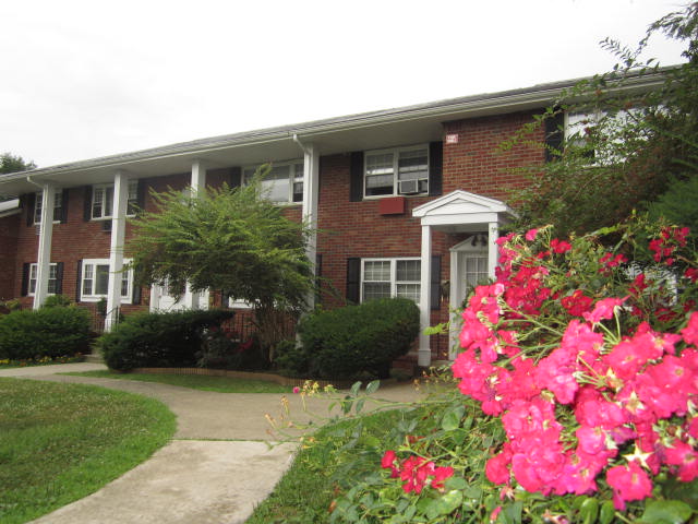 Knox Village Apartments, exterior, red brick building, 2 levels, white covered entrance, grass, trees, bushes, pink flowers