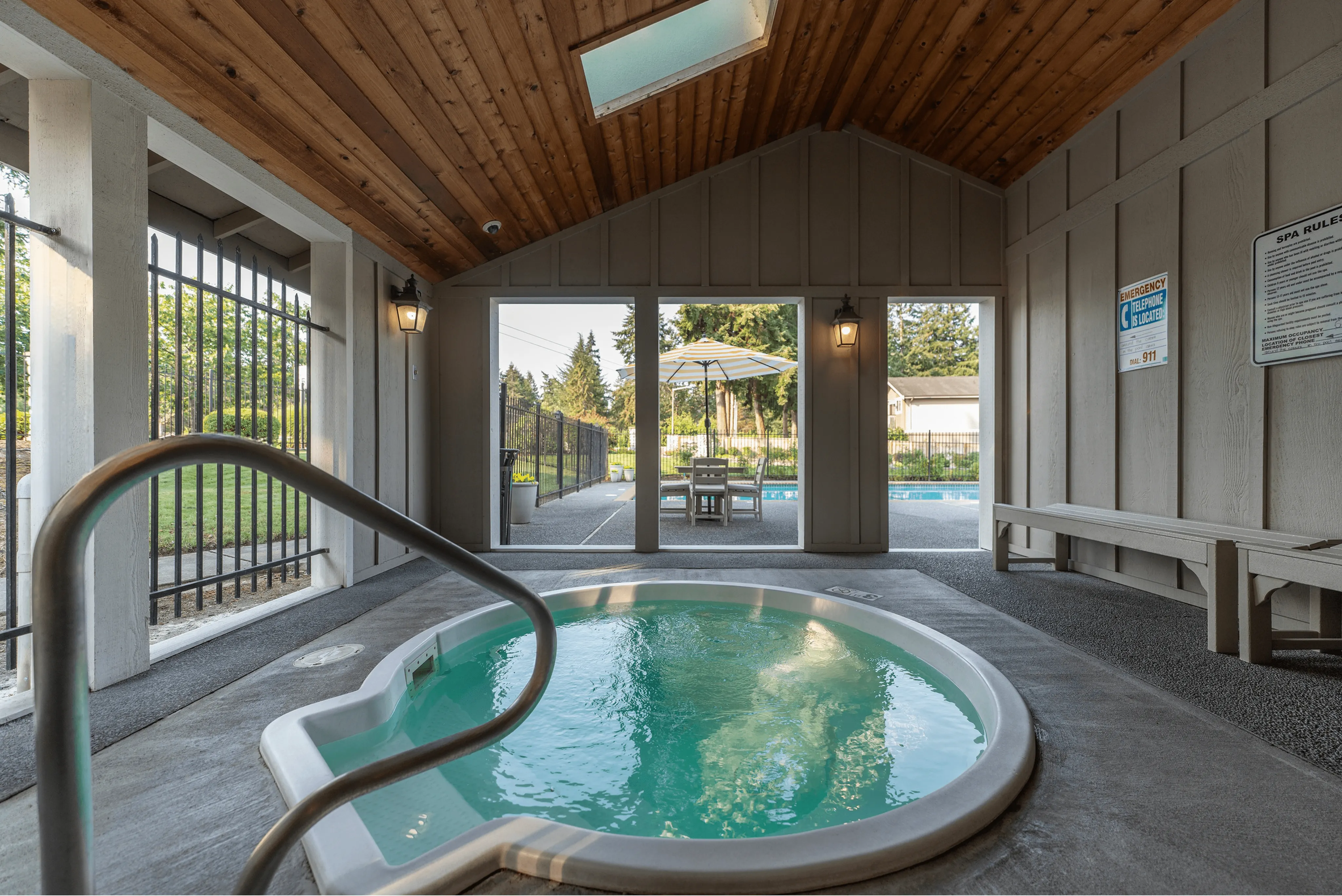 Inviting spa area with hot tub under a wooden ceiling, opens to a sunlit outdoor pool area with seating.