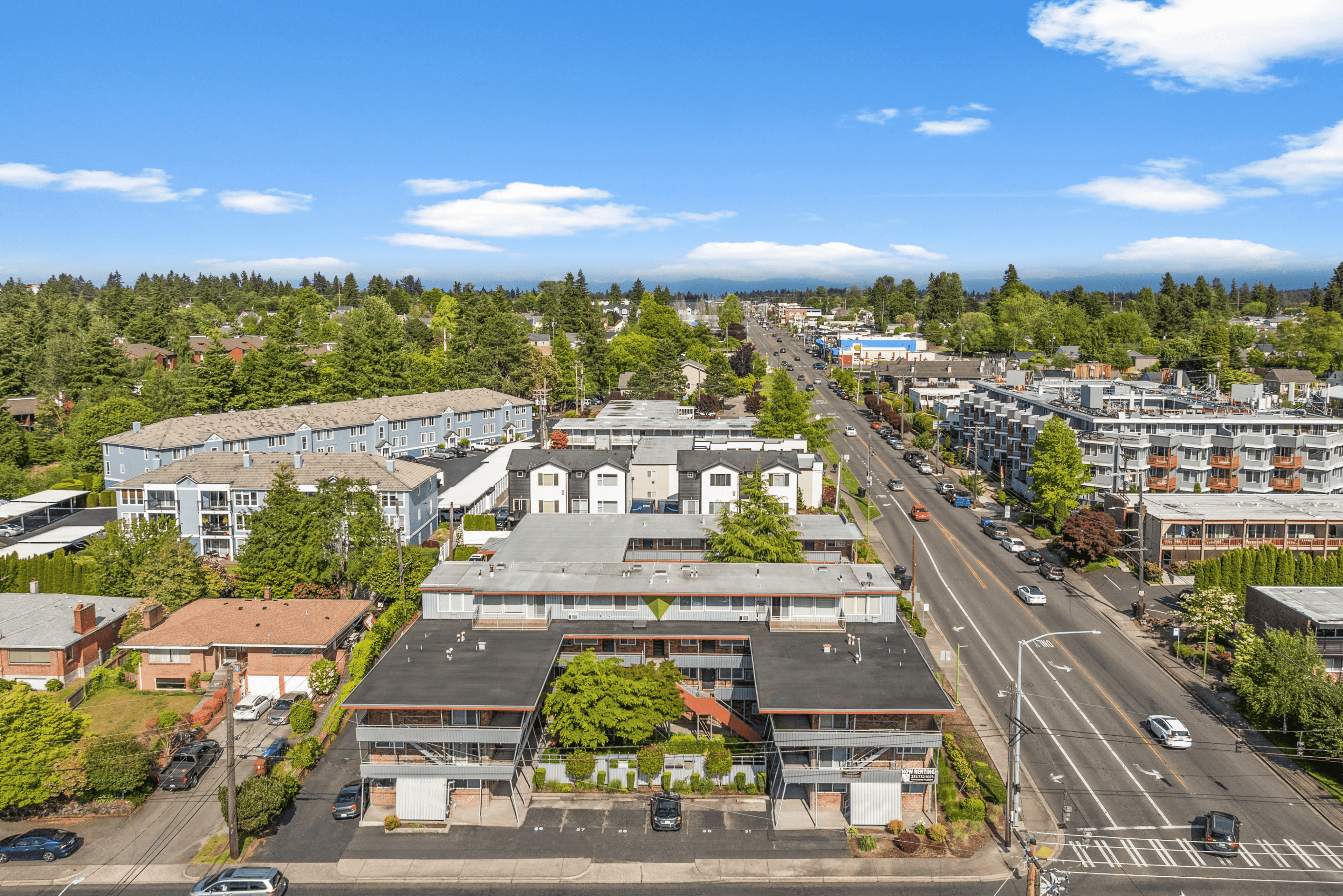 Aerial view of a vibrant neighborhood featuring modern apartment complexes surrounded by lush greenery and a bustling main road under a clear blue sky.
