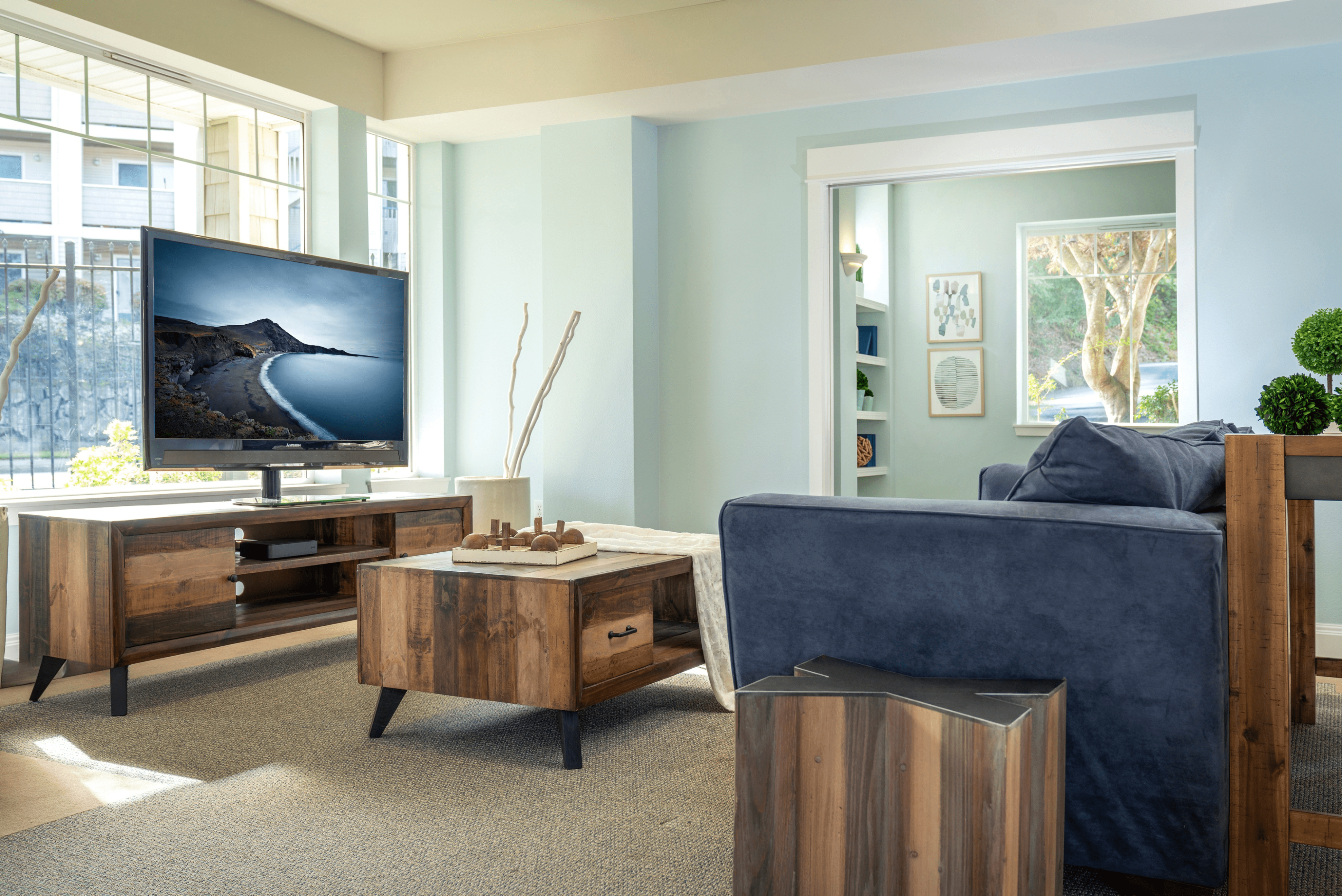 Inviting living room with modern design, featuring a plush blue sofa, rustic wood furniture, large windows, and a sleek flat-screen TV.