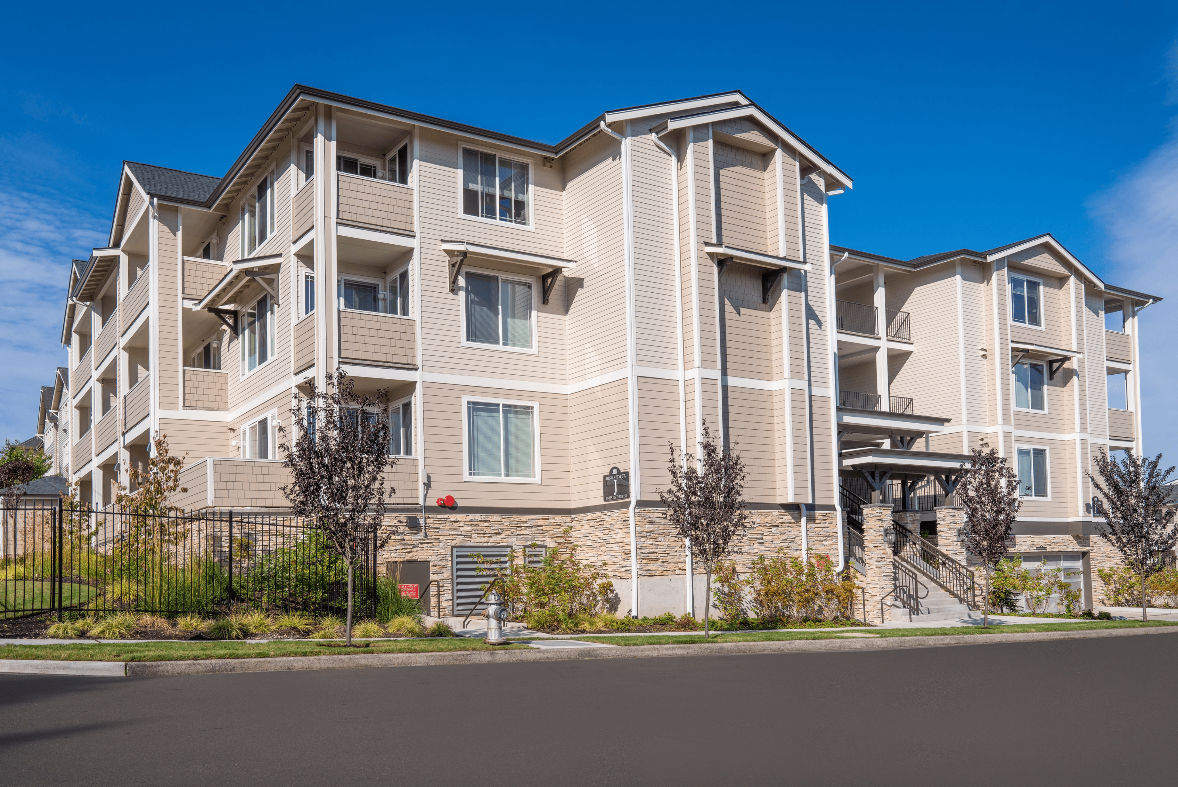 Modern three-story apartment building with balconies and lush landscaping under a clear blue sky.