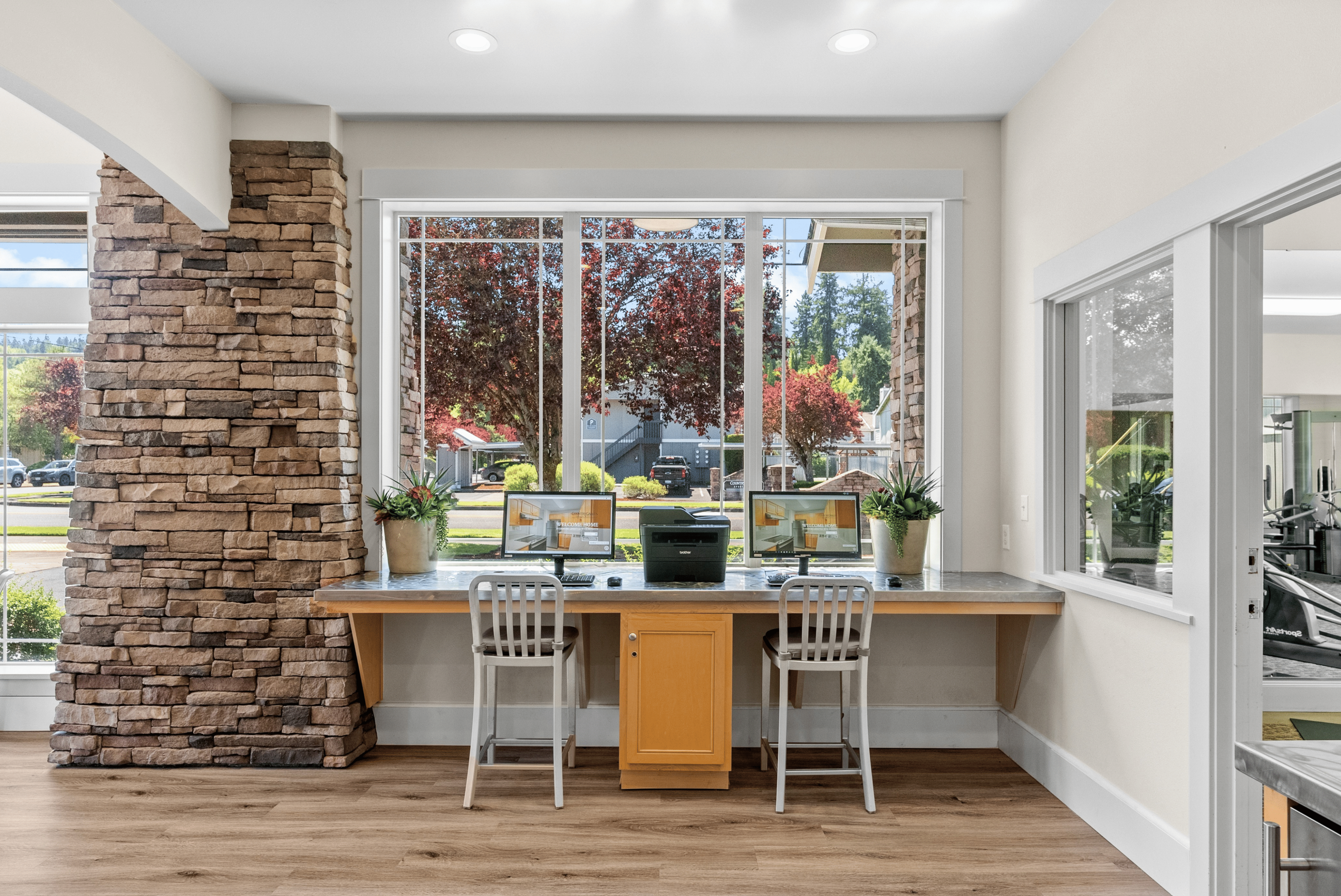 Modern communal workspace with large windows, featuring two computer stations, stylish chairs, and beautiful garden views.
