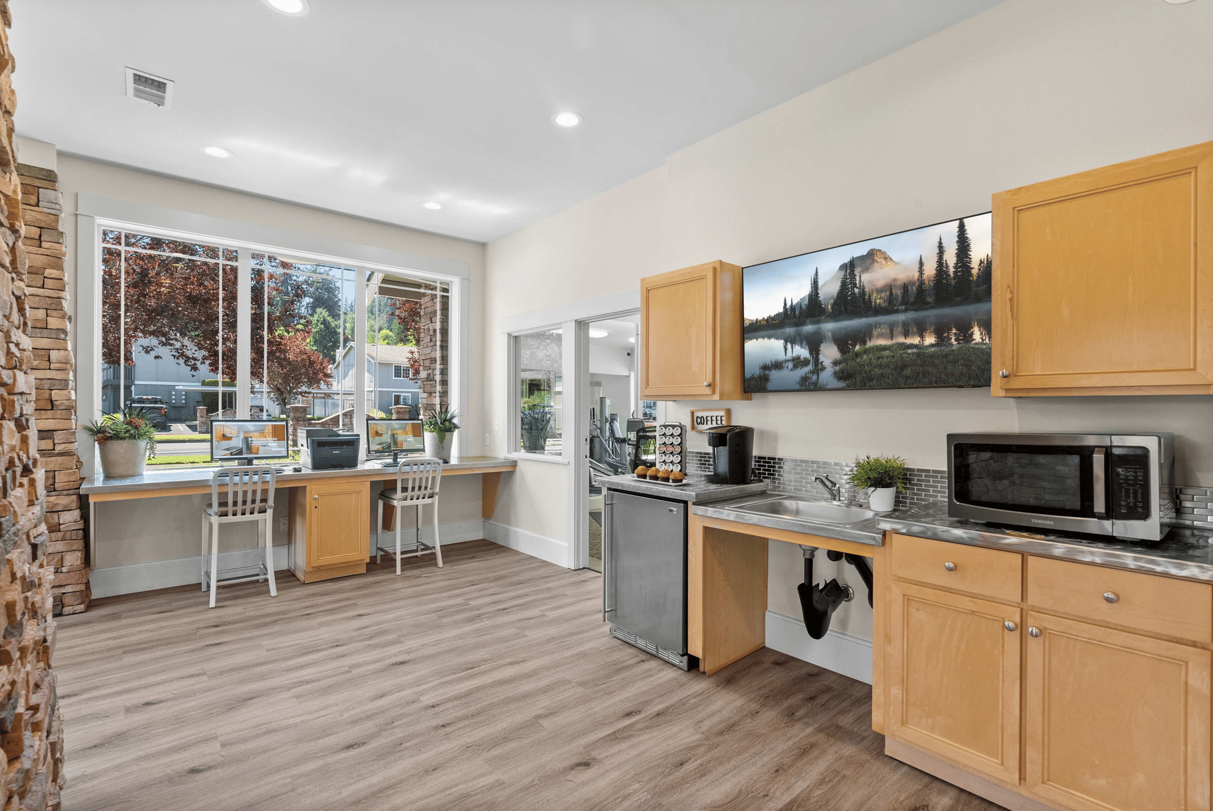 Inviting workspace with large windows and modern kitchen setup, featuring sleek cabinetry, wooden flooring, and stylish decor.
