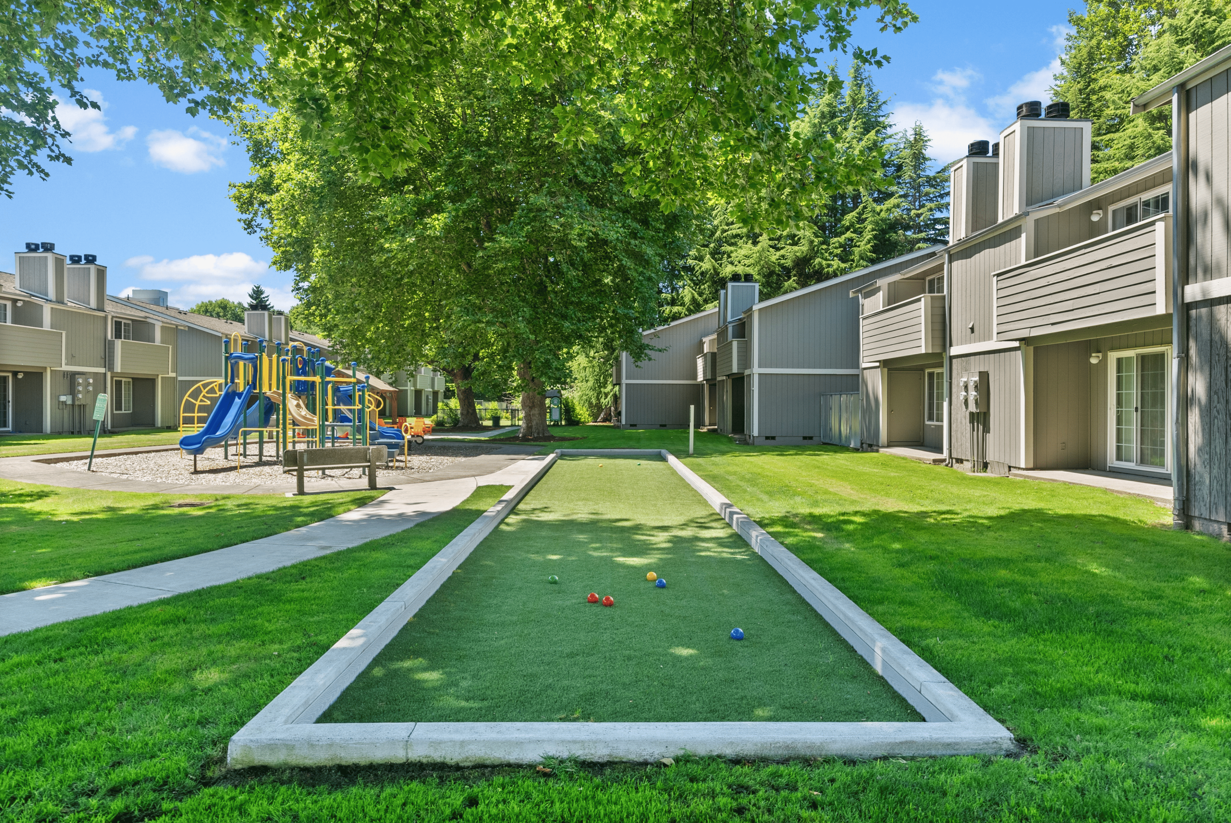 Charming apartment exterior with a playground and bocce ball court surrounded by lush greenery.