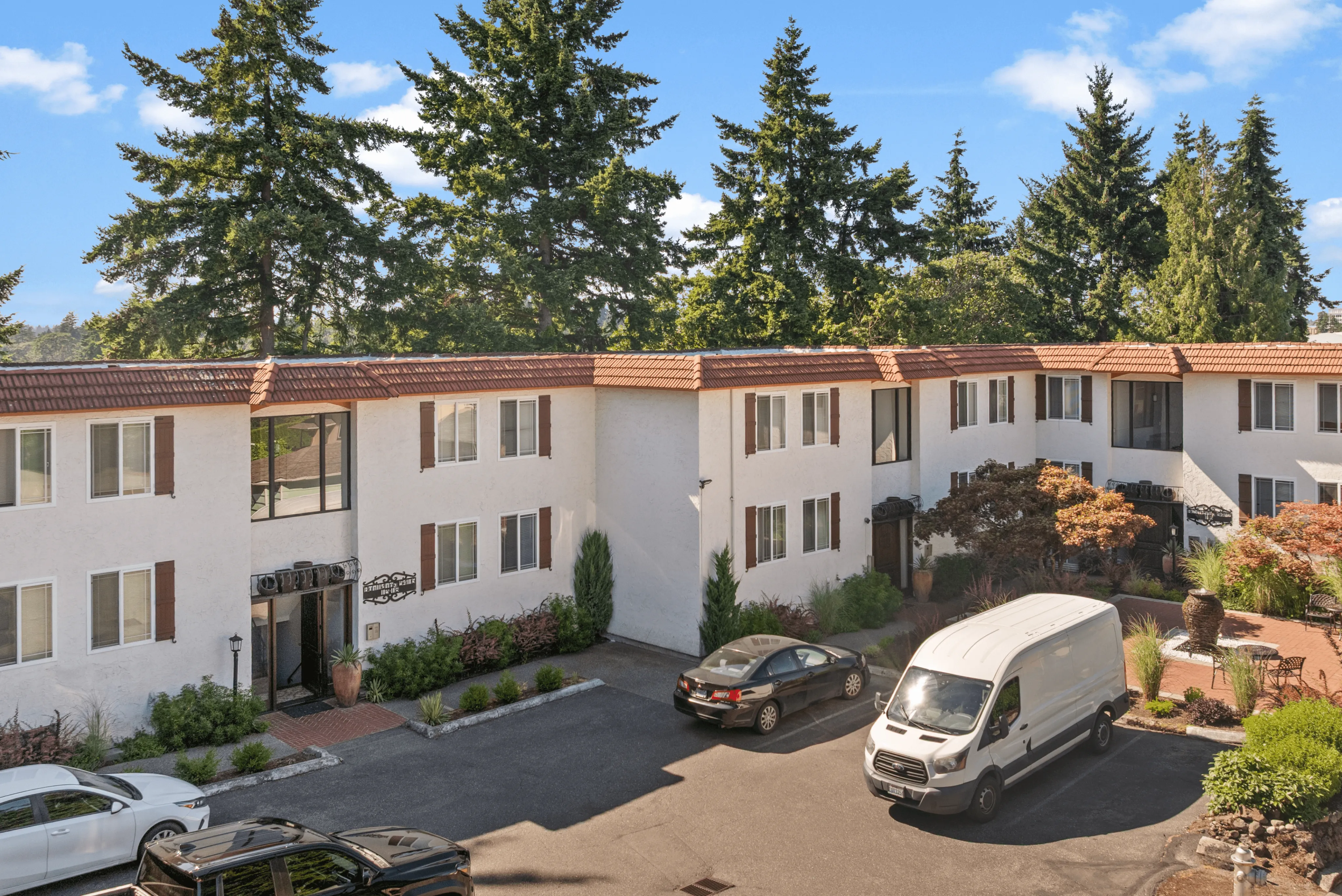 Charming two-story apartment complex with red-tiled roof, surrounded by lush greenery and ample parking space under a clear blue sky.
