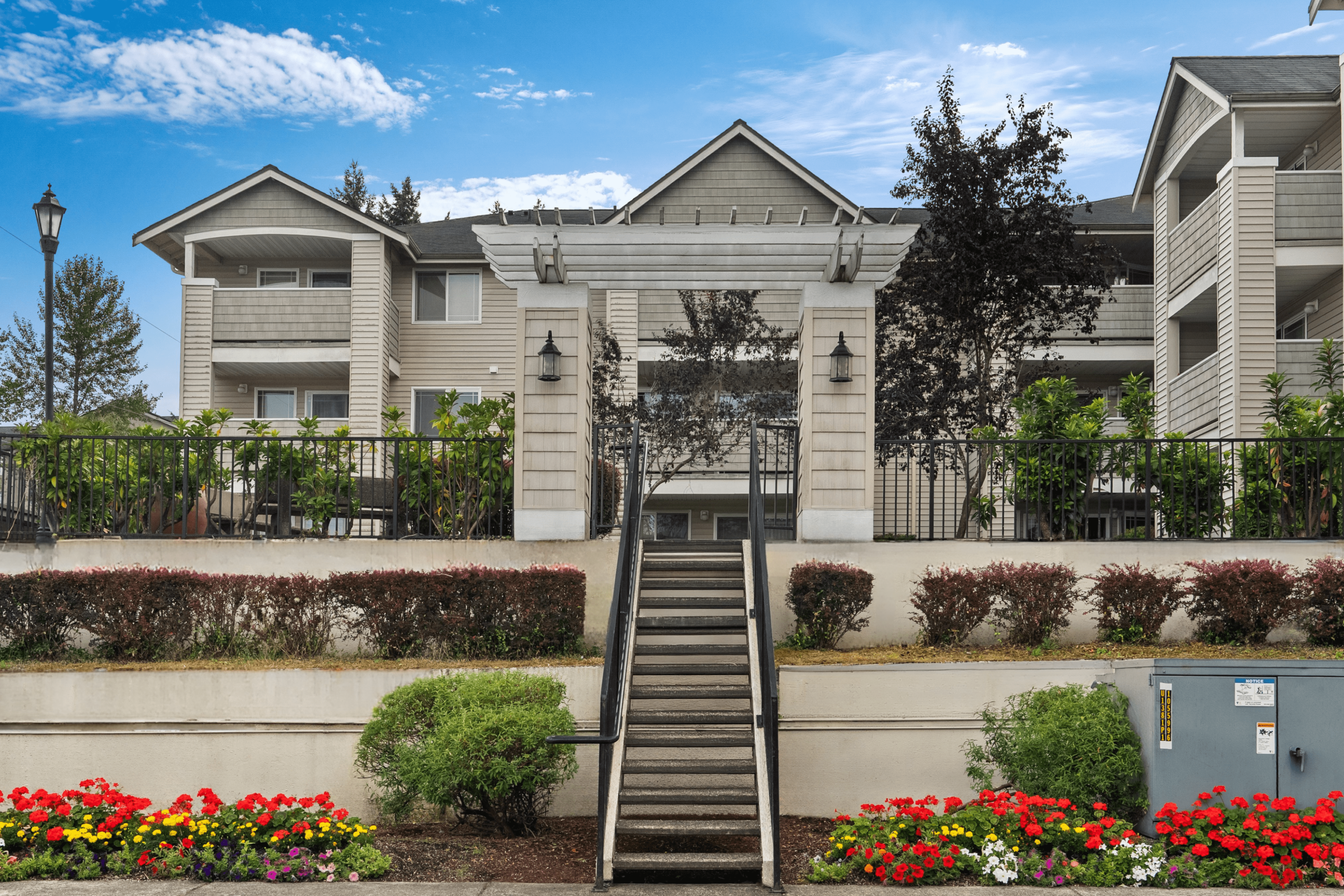 Inviting facade of a modern apartment complex with lush landscaping, vibrant flower beds, and classic architectural details under a clear blue sky.