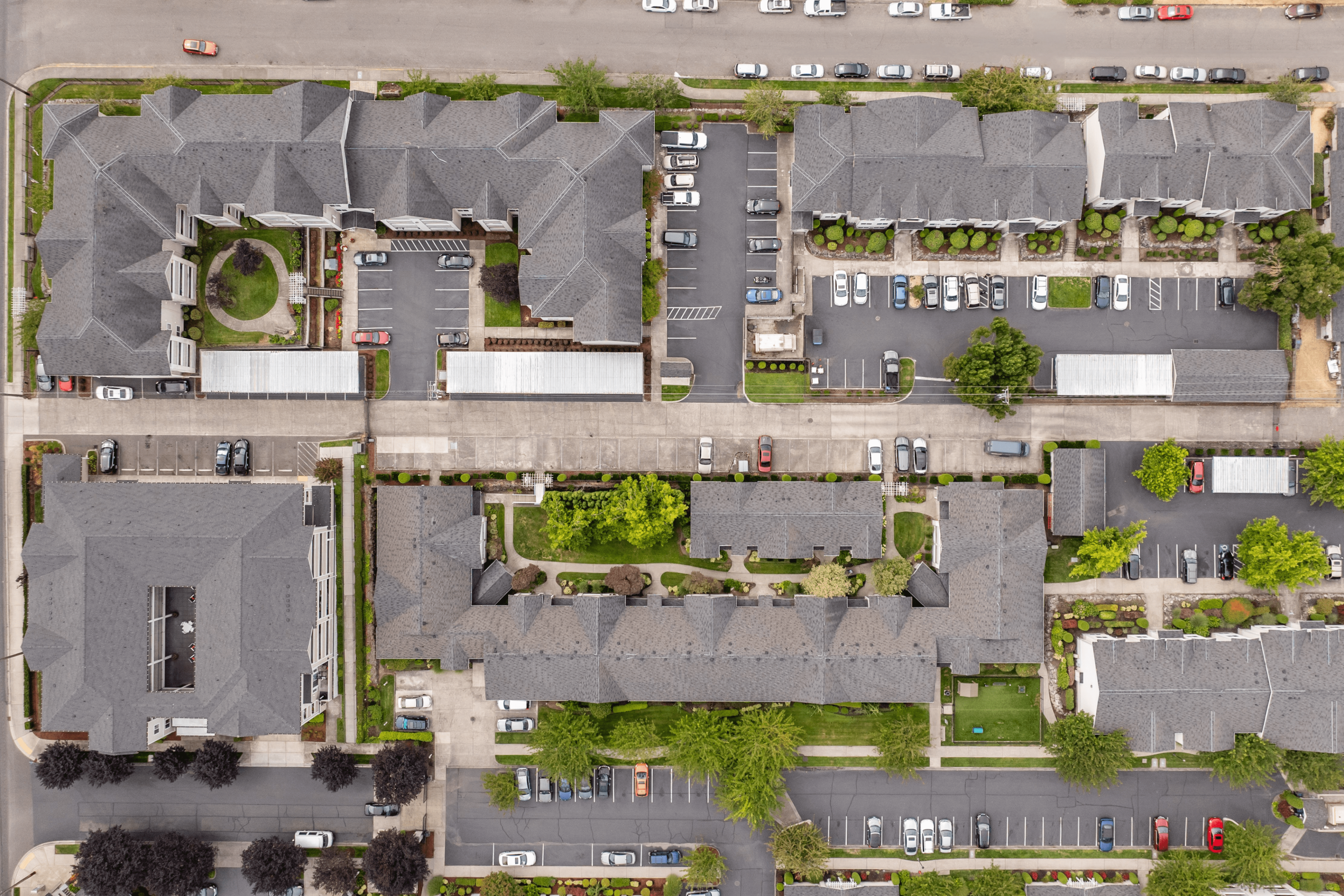 Aerial view of a charming apartment complex featuring multiple residential buildings, neatly arranged pathways, and ample parking space, surrounded by lush green trees and landscaping.