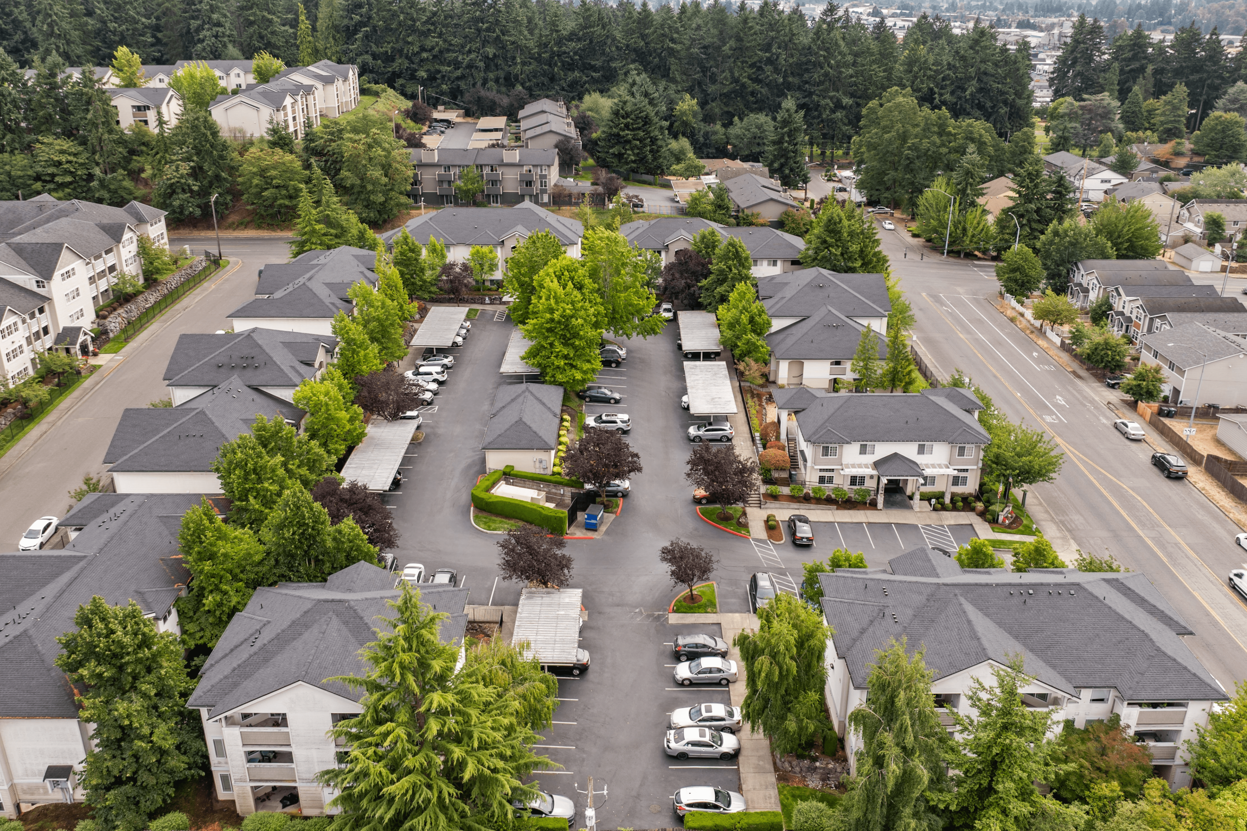 Charming aerial view of a serene apartment complex surrounded by lush greenery, showcasing ample parking and neatly arranged residential buildings.