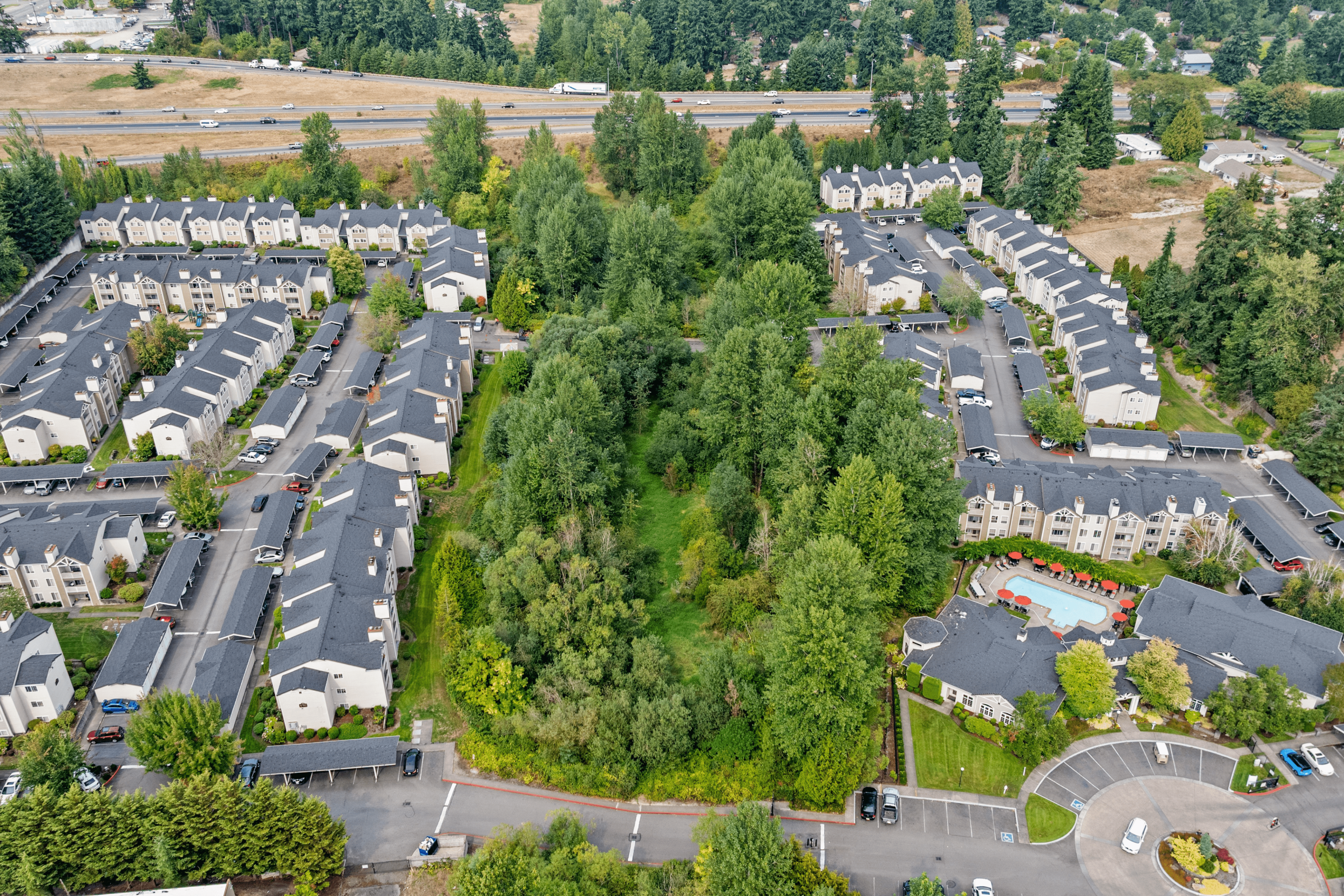 Aerial view of a charming apartment complex surrounded by lush greenery and landscaped gardens, featuring inviting pathways and a pristine community swimming pool with red umbrellas.
