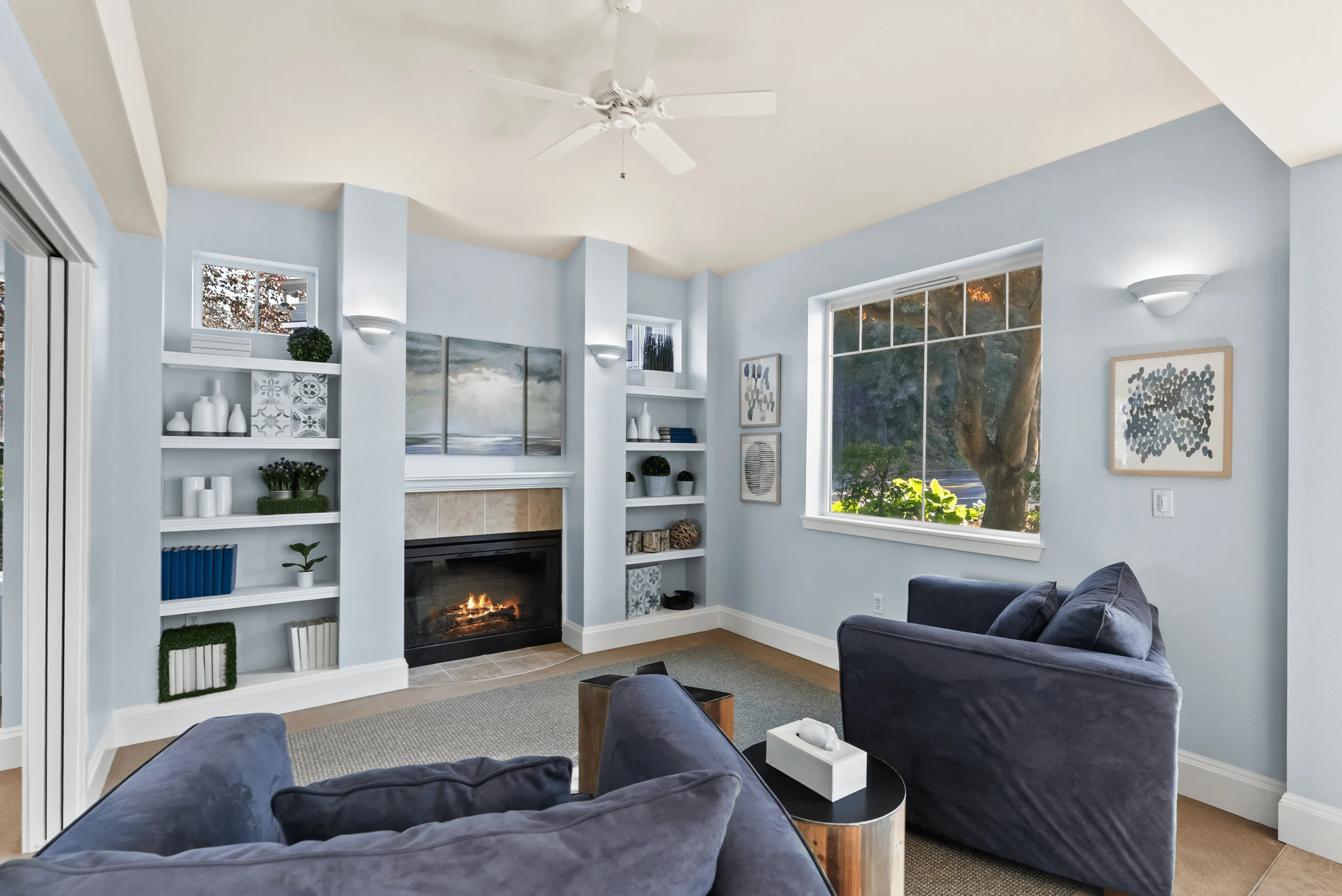 Inviting living room with elegant built-in shelves, cozy fireplace, modern blue couches, and large window with lush garden view.