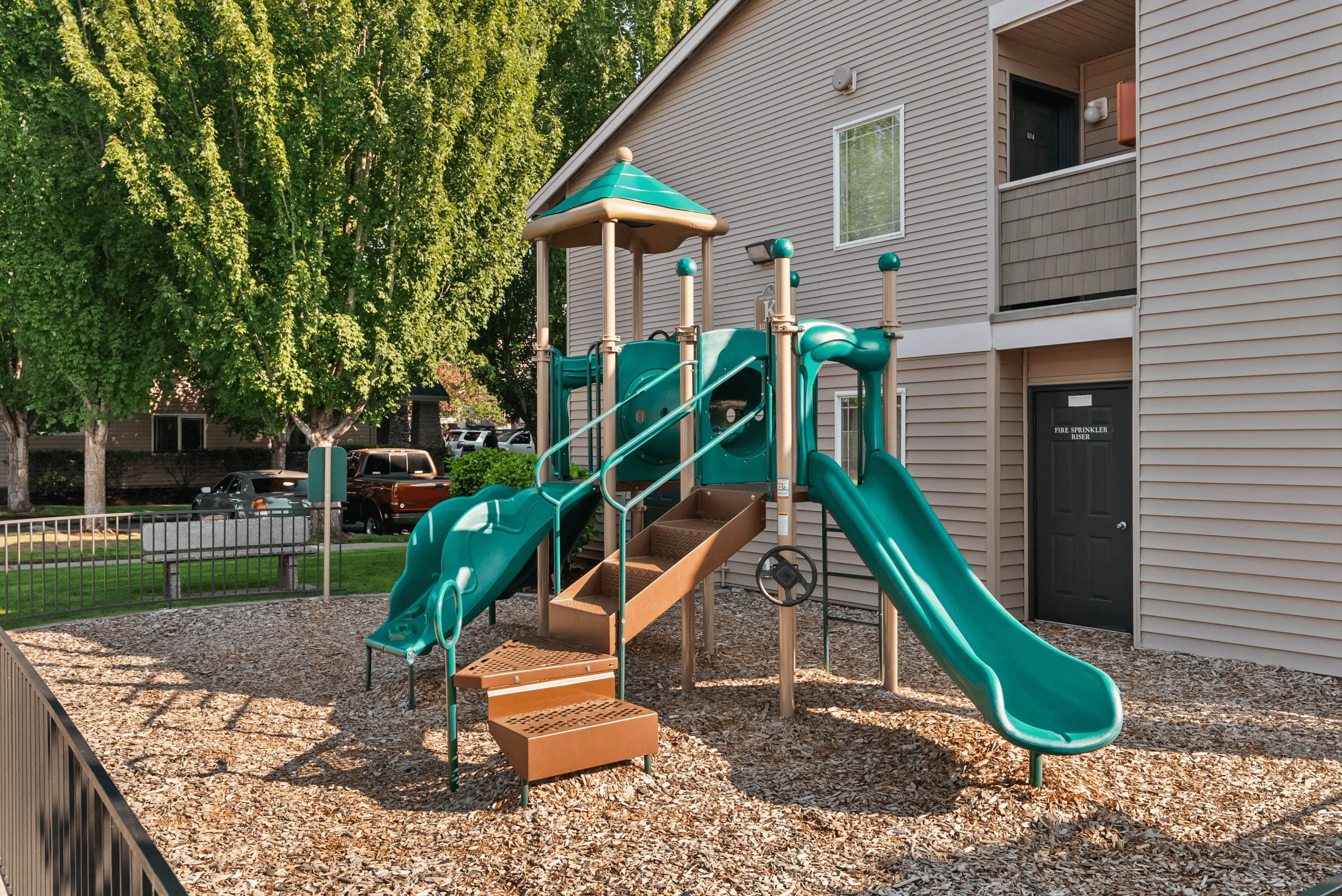 Charming apartment playground featuring vibrant green slides and shaded play structure, nestled in a serene community setting with lush trees.