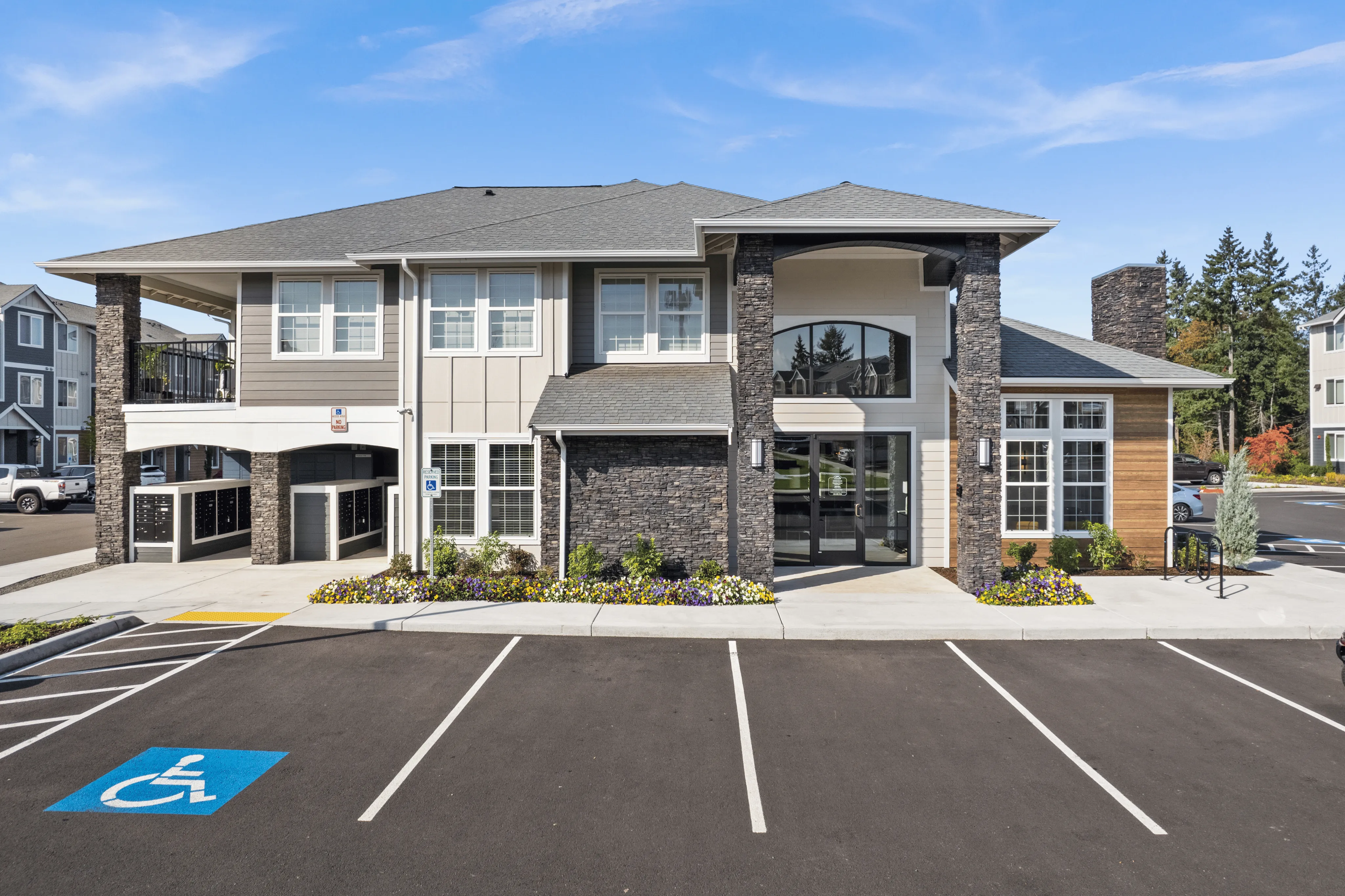 Modern apartment complex entrance featuring stone accents, large windows, and well-kept landscaping under a clear blue sky.