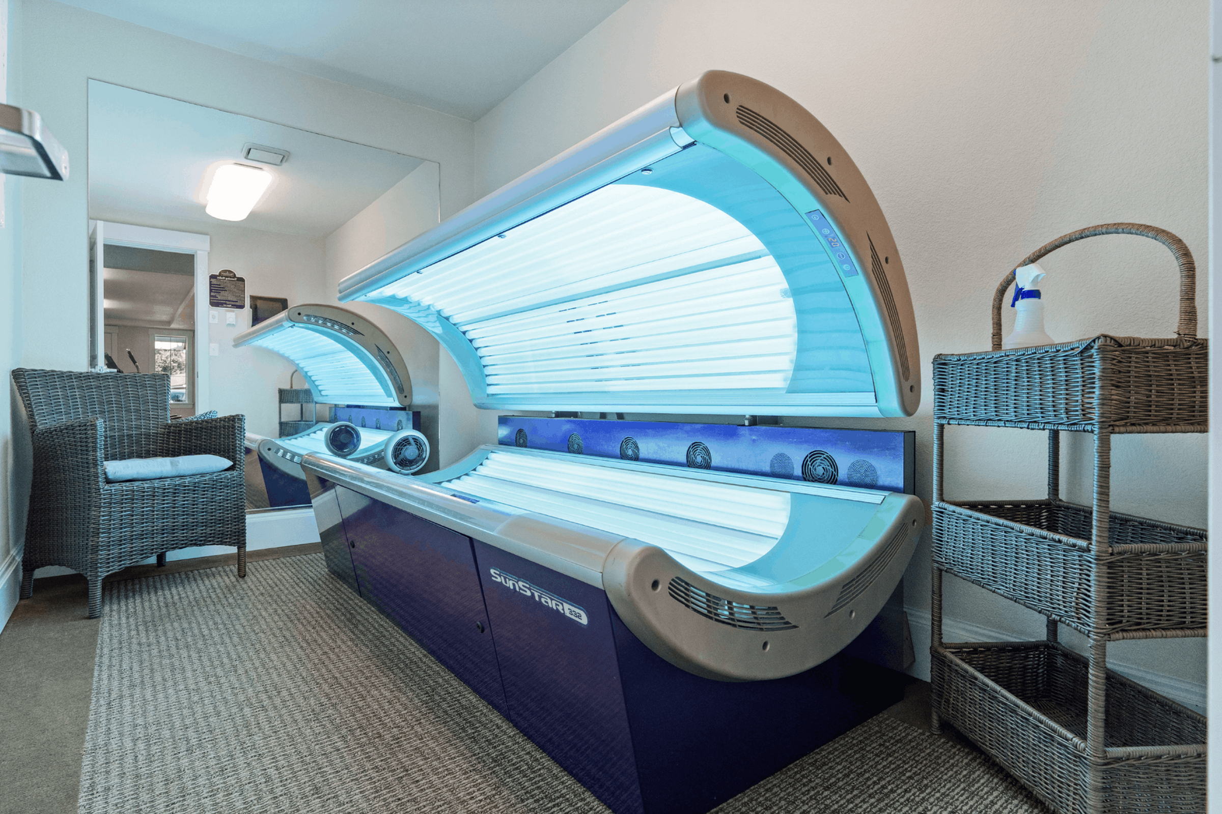 Modern indoor tanning room with sleek SunStar tanning beds, stylish wicker chair, and organized basket storage.