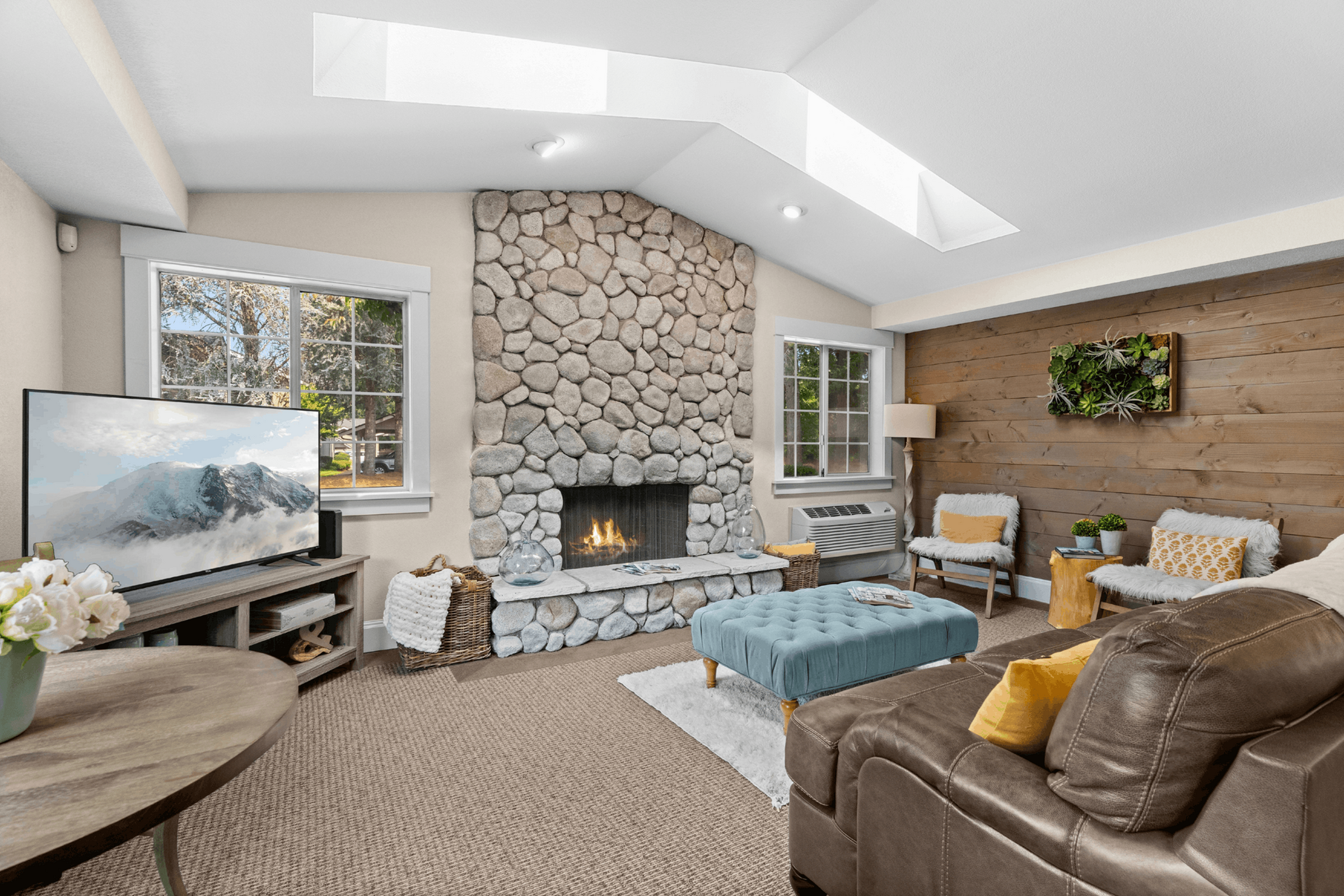 Cozy living room with a rustic stone fireplace, large windows, and modern decor. Features a comfortable seating area with a stylish blue ottoman and a lush indoor plant wall accent.