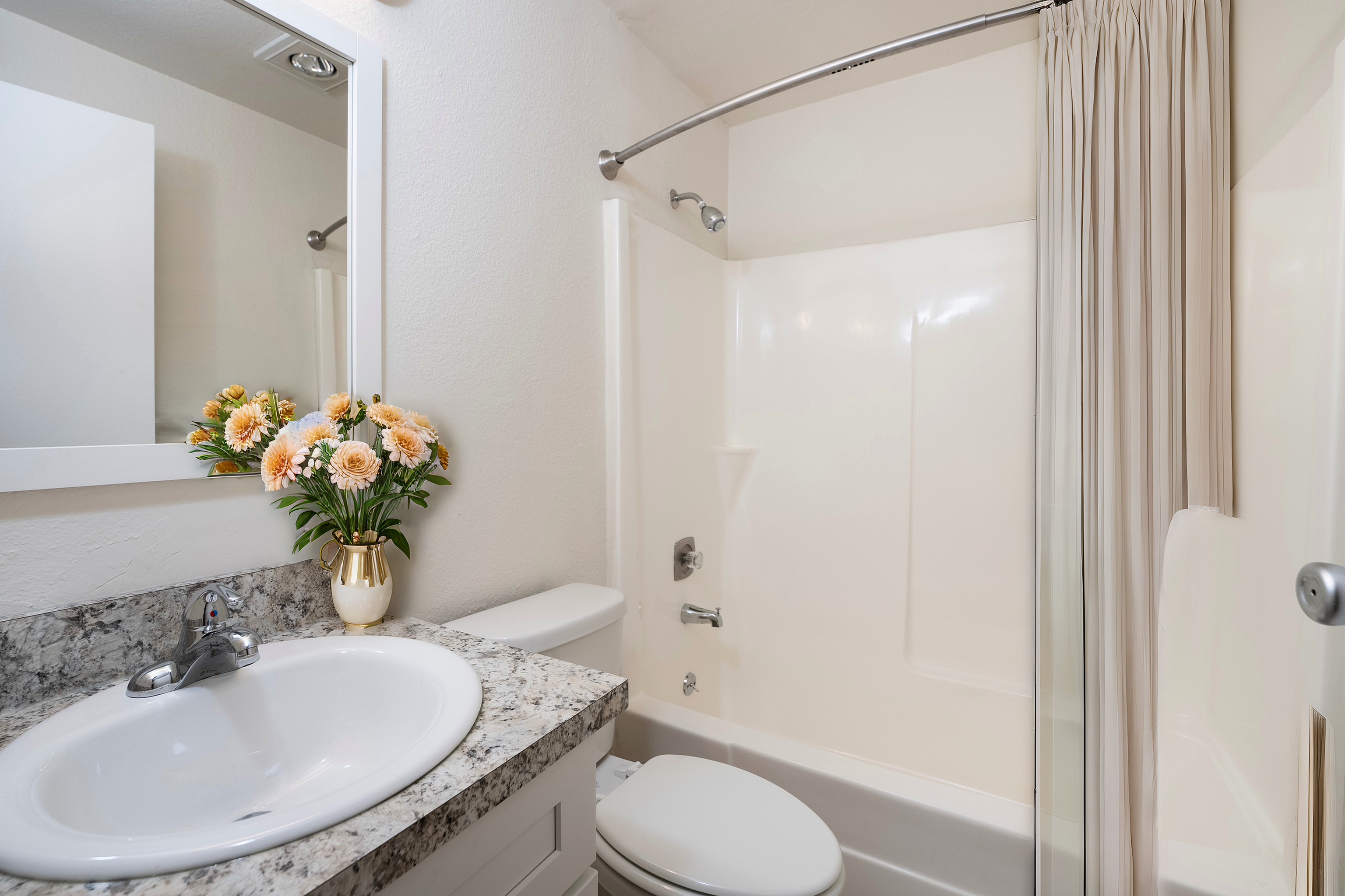 Bright bathroom with a granite countertop and floral arrangements beside a shower and toilet.