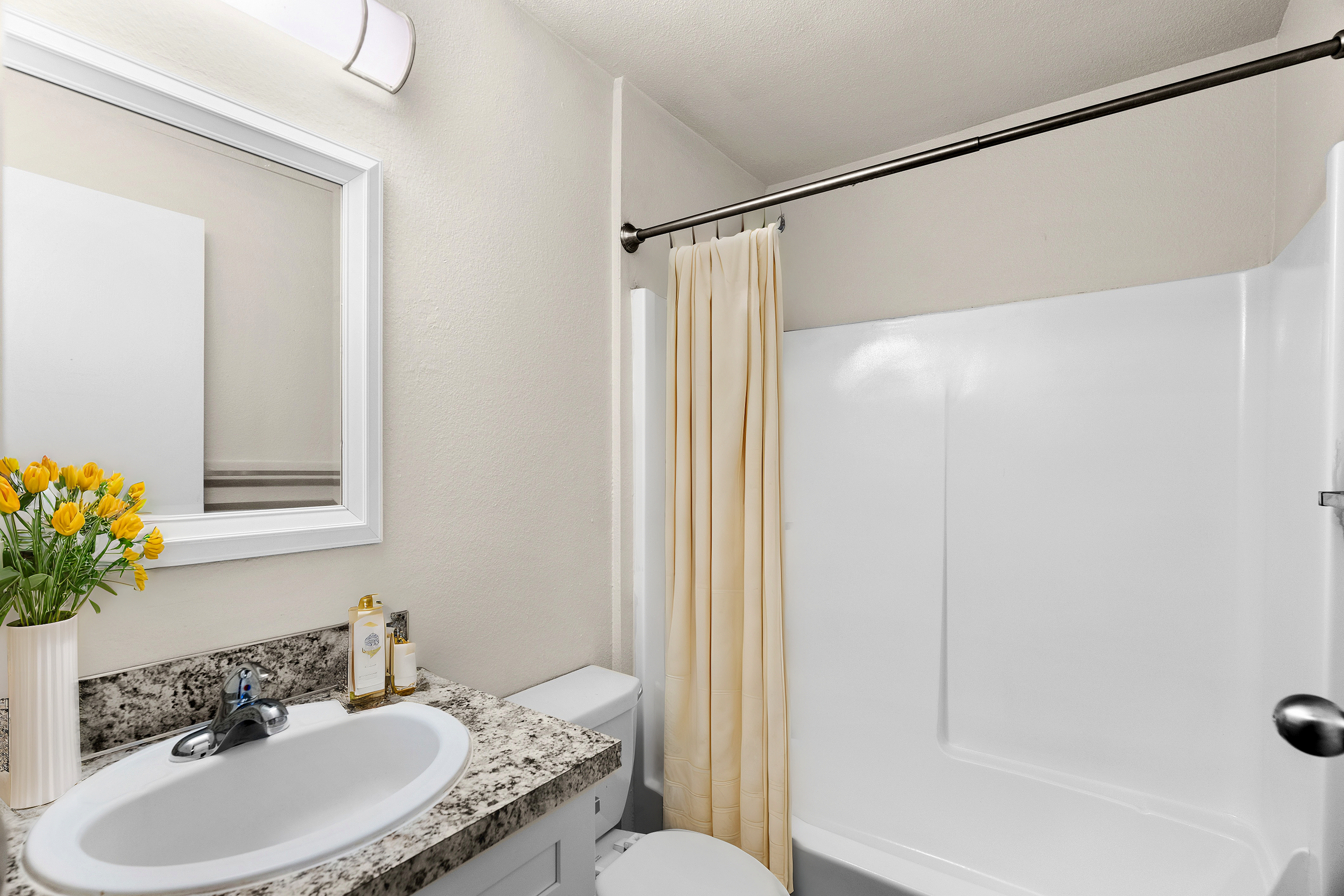 Bright and modern bathroom featuring a sleek countertop, vase with yellow flowers, and a bathtub with a cream-colored shower curtain.