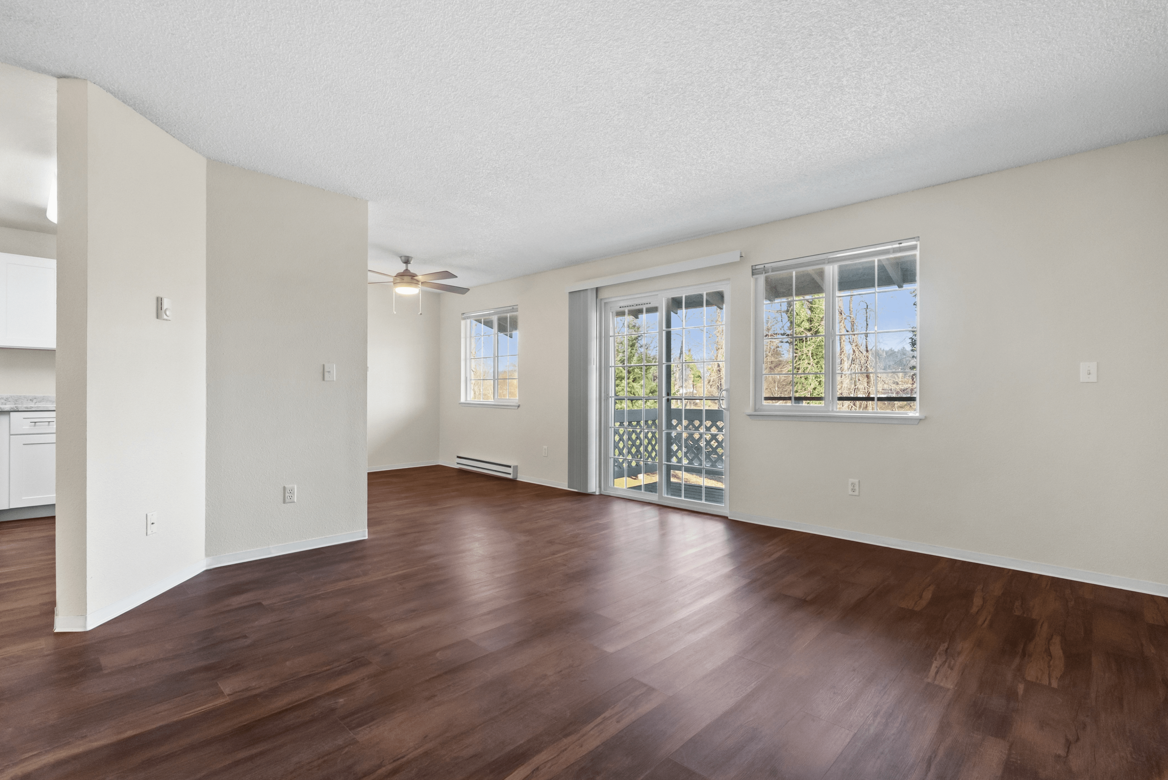 Sunlit living room with wooden flooring, large windows, and sliding glass doors leading to a balcony.