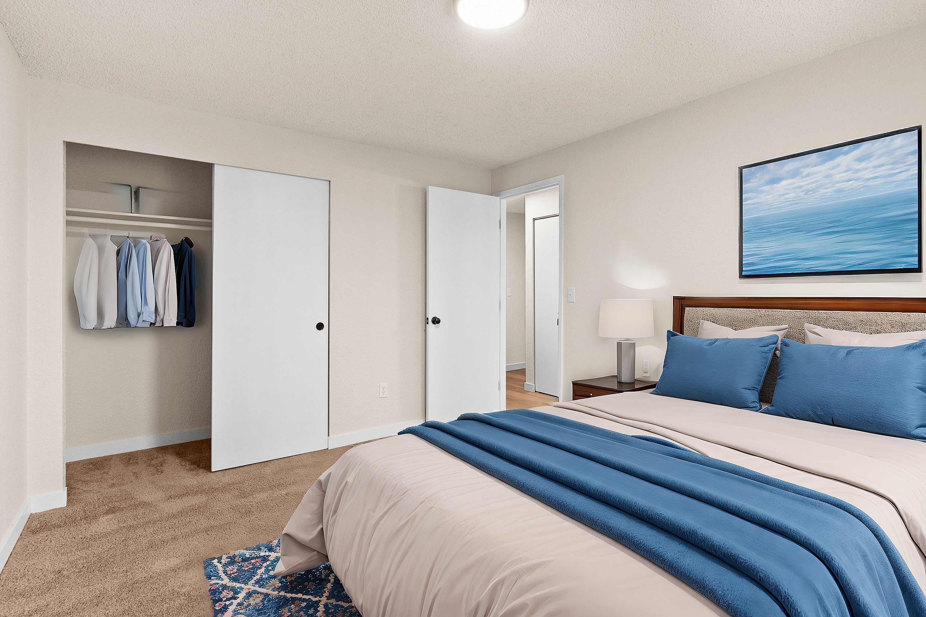 Inviting bedroom featuring a well-made bed with blue accents, a closet with clothes, and a serene ocean painting.
