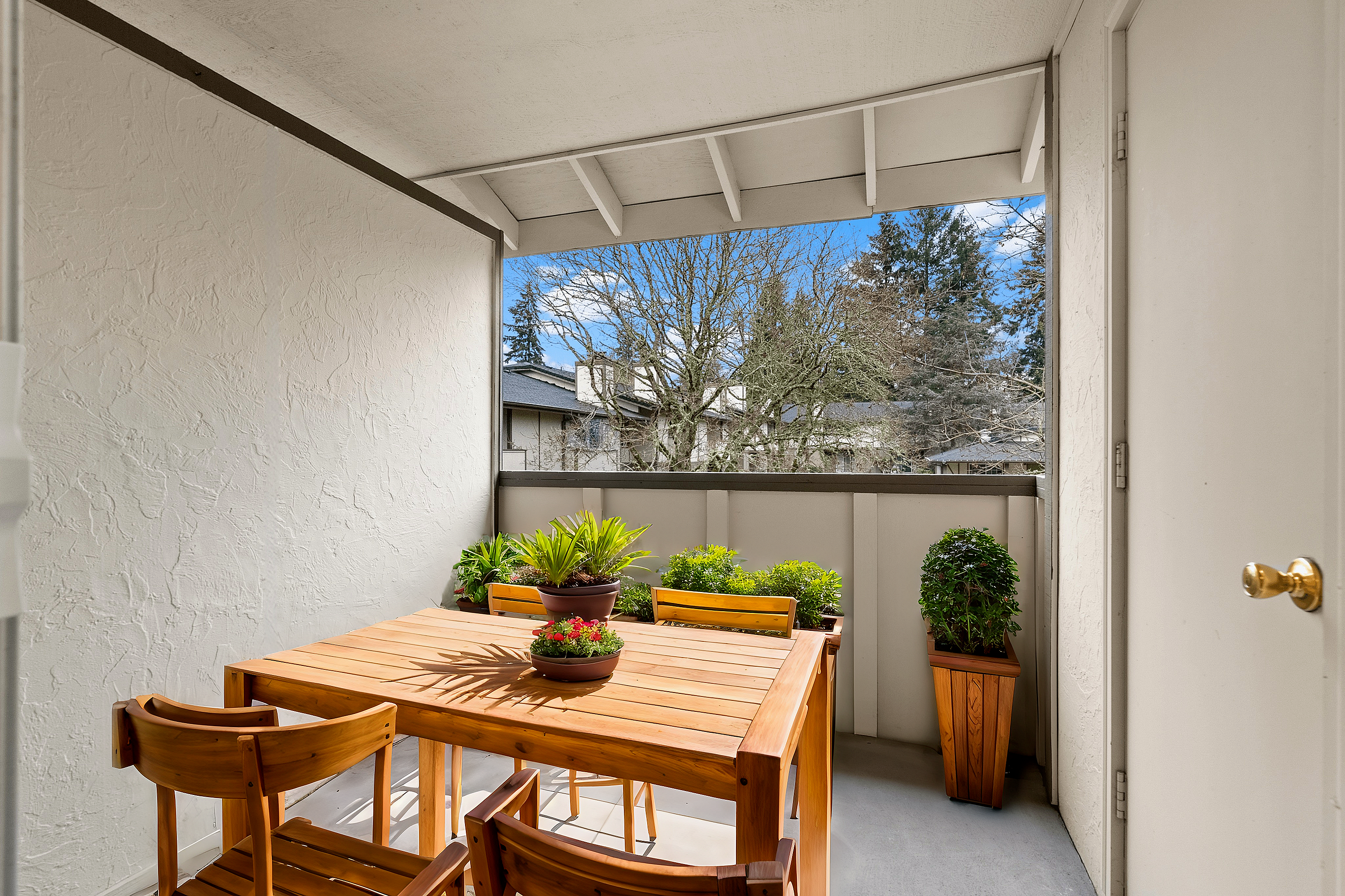 Cozy balcony with wooden table and chairs, surrounded by potted plants and a view of trees.