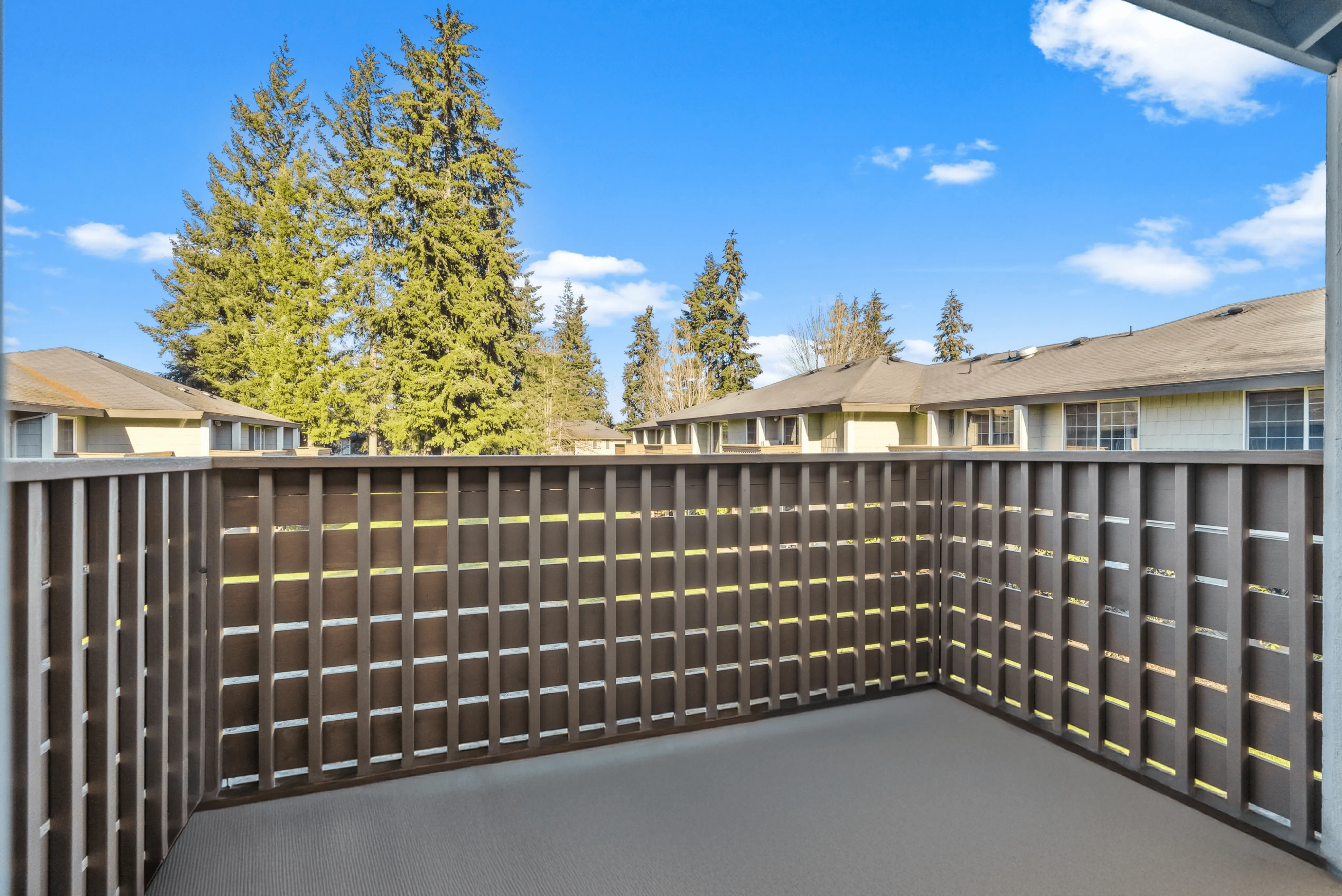 Cozy balcony with lattice railing overlooking lush evergreen trees and neighboring townhomes under a blue sky.