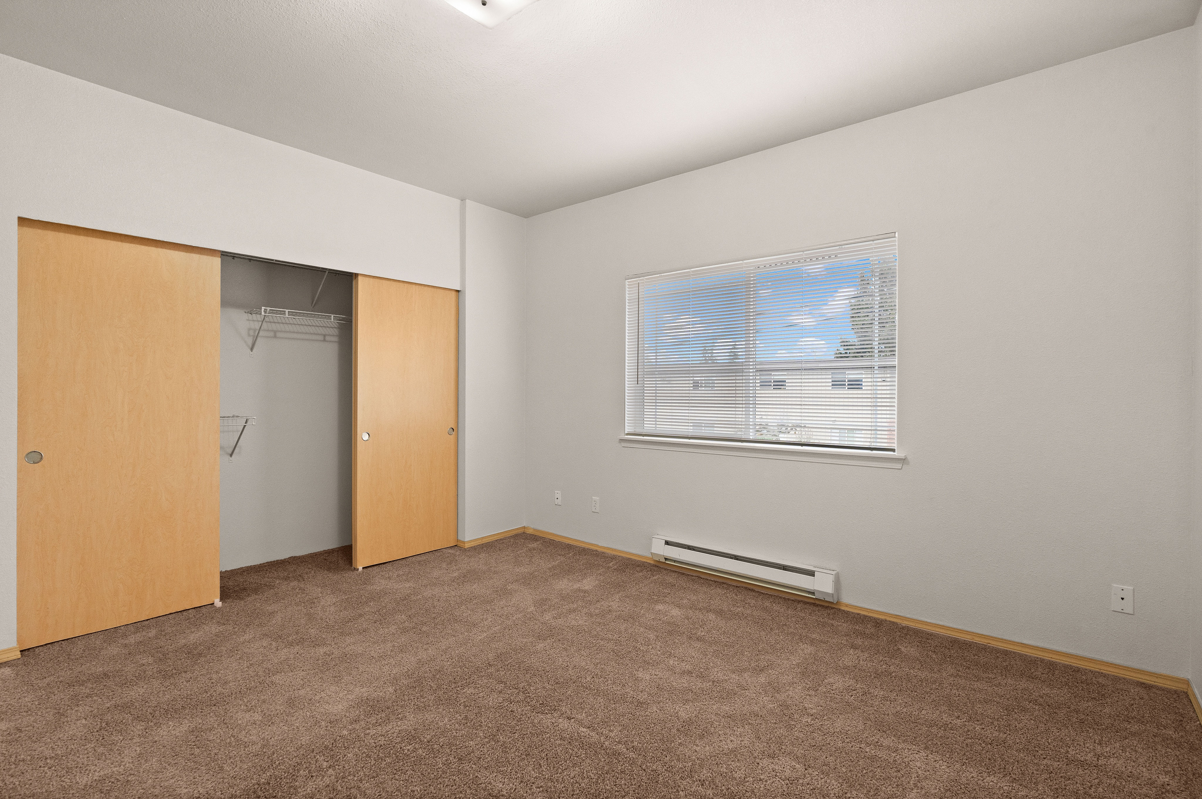 Bright and spacious bedroom with plush carpet, large window with blinds, and ample closet space with sliding wooden doors.