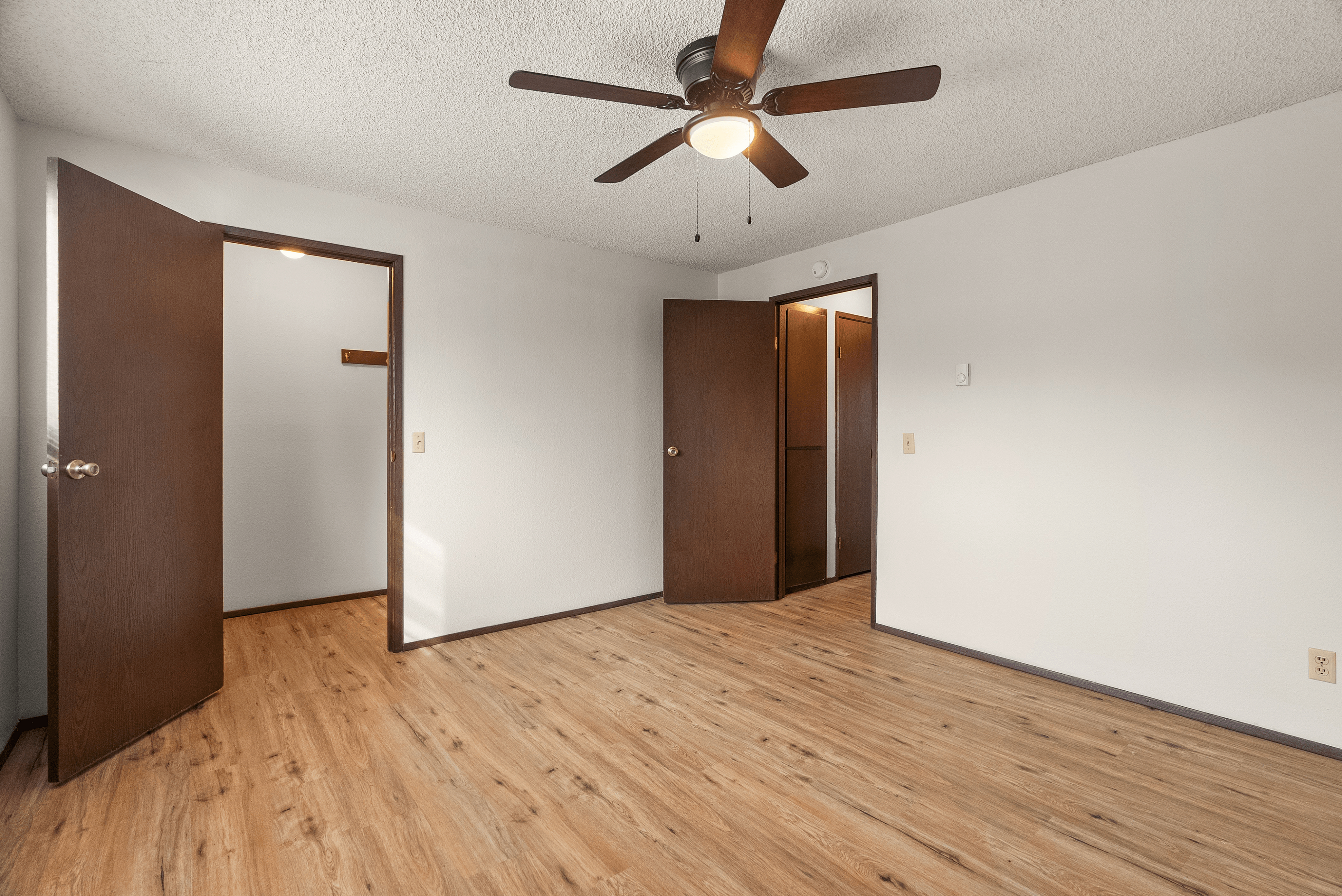 Spacious bedroom with elegant wood flooring, white walls, and a stylish ceiling fan offering a warm and inviting atmosphere.