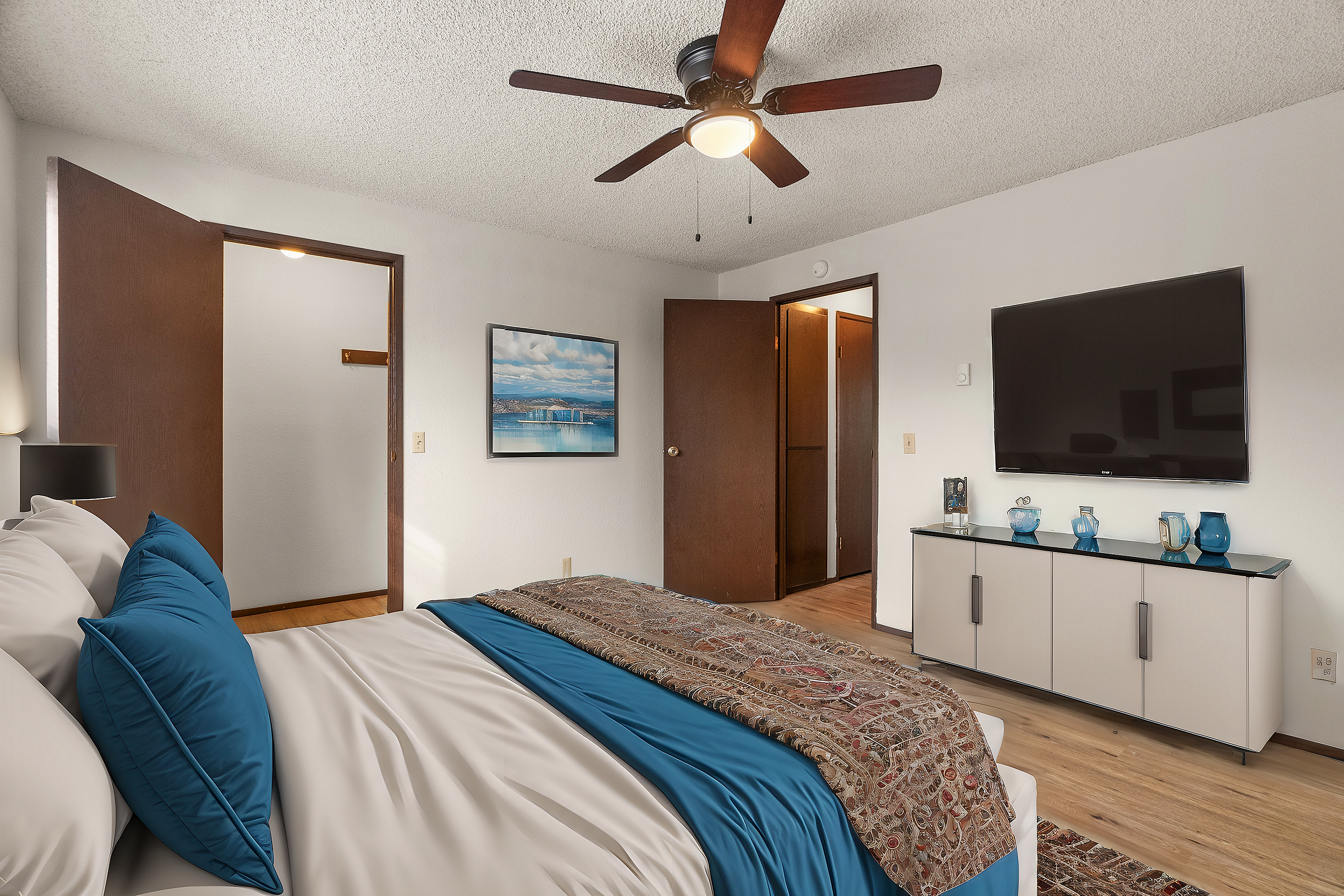 Cozy bedroom with modern decor, featuring a ceiling fan, vibrant bedding, wall-mounted TV, and elegant hardwood flooring.