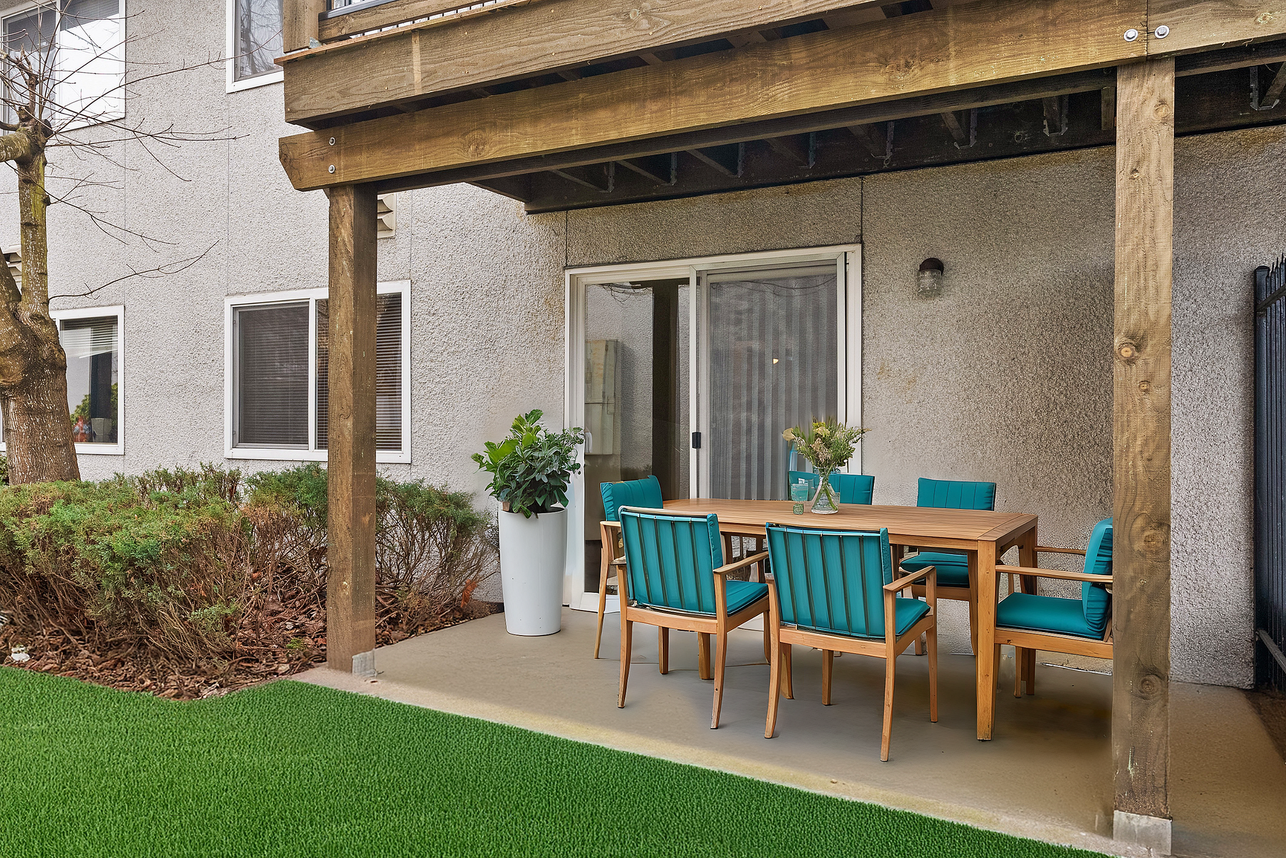 Charming outdoor patio with a wooden dining set and turquoise cushions, surrounded by lush greenery and modern architecture.