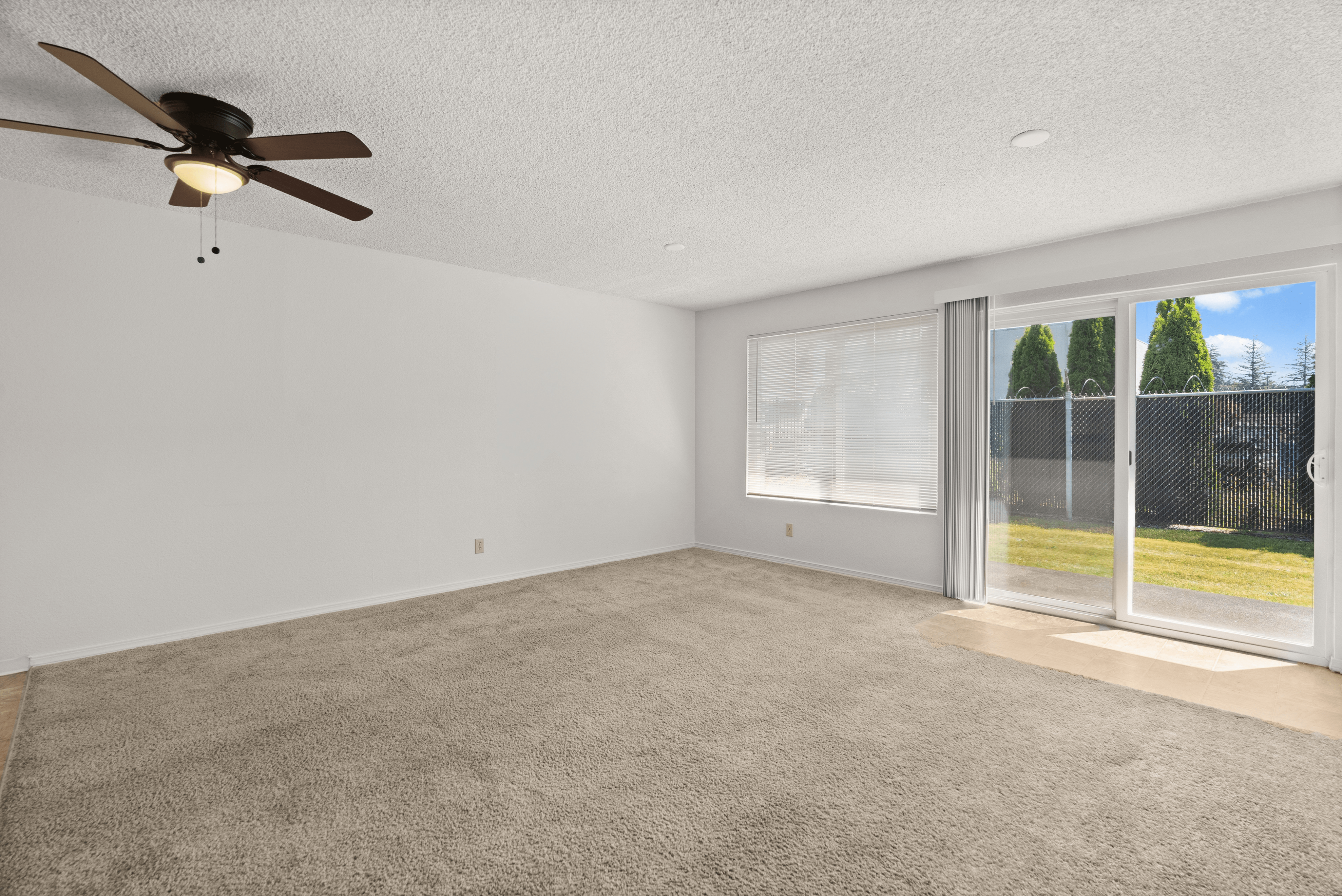 Bright and airy living room with plush carpet, ceiling fan, and large sliding glass door leading to a lush green backyard.