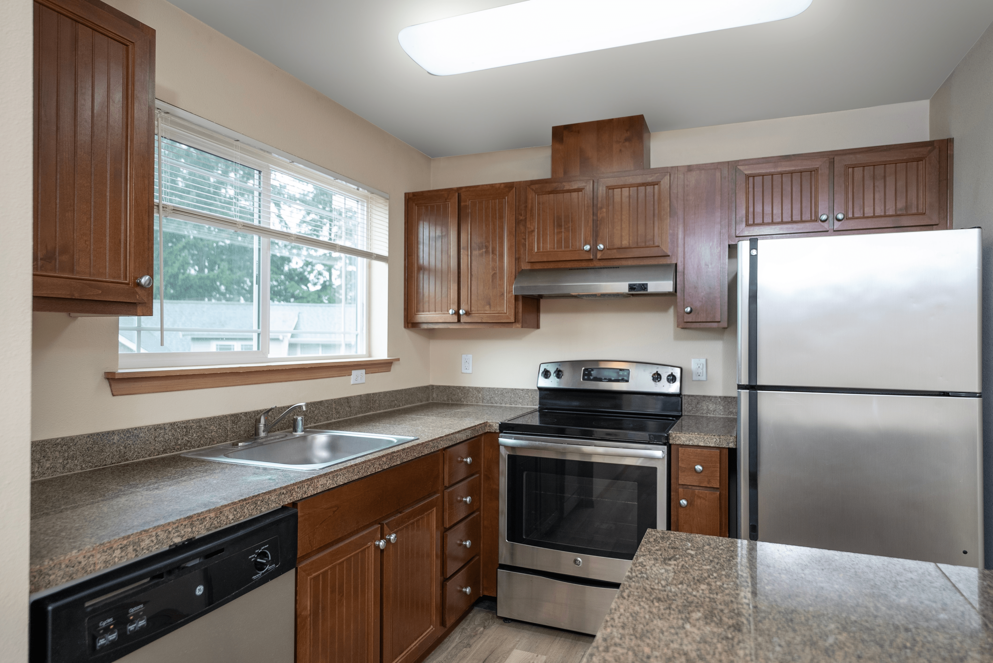 Modern kitchen with stainless steel appliances, wood cabinetry, and granite-style countertops. A large window allows natural light to brighten the space.