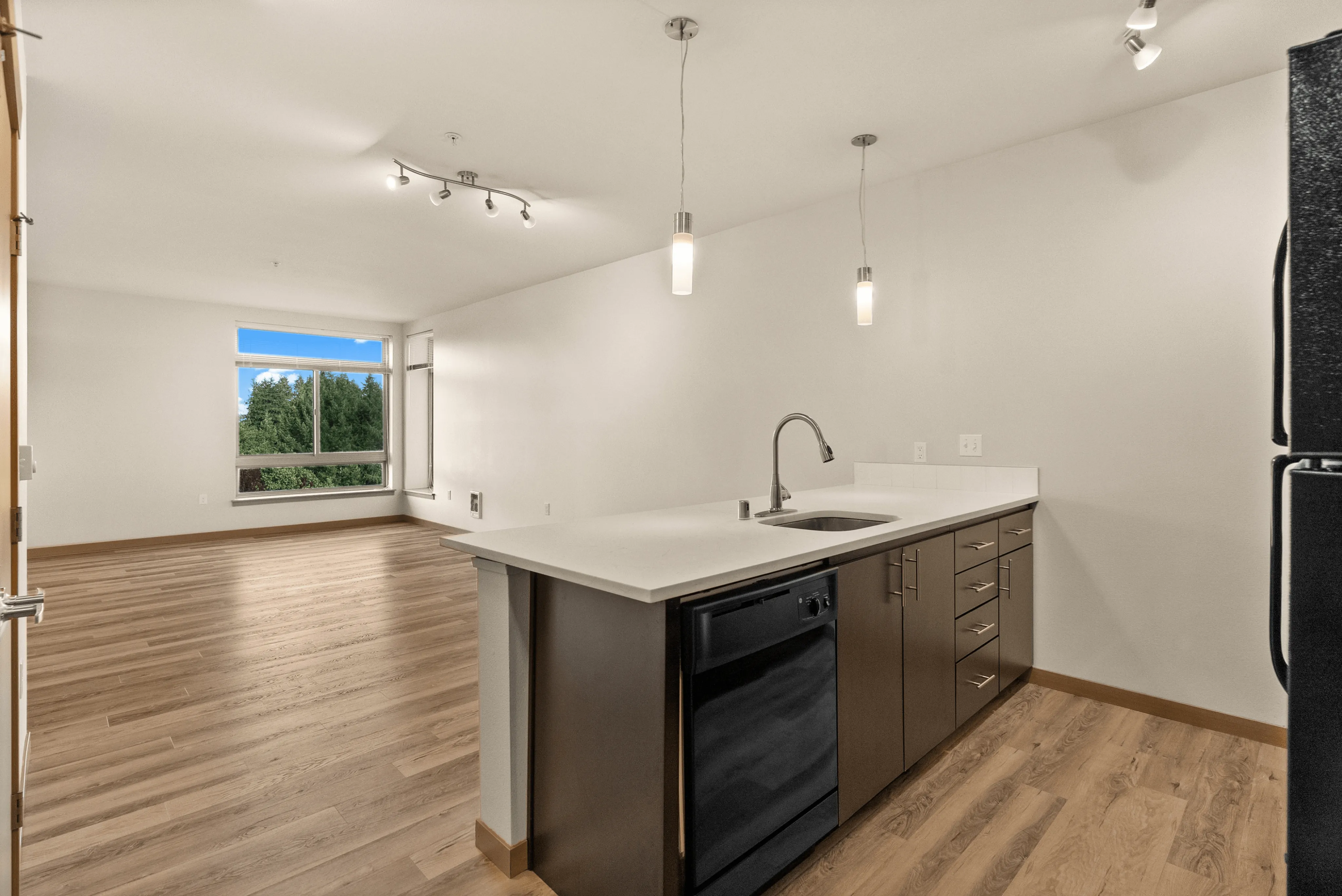 Open-concept apartment interior with a kitchen island, pendant lighting, and large living area with a picture window overlooking greenery.