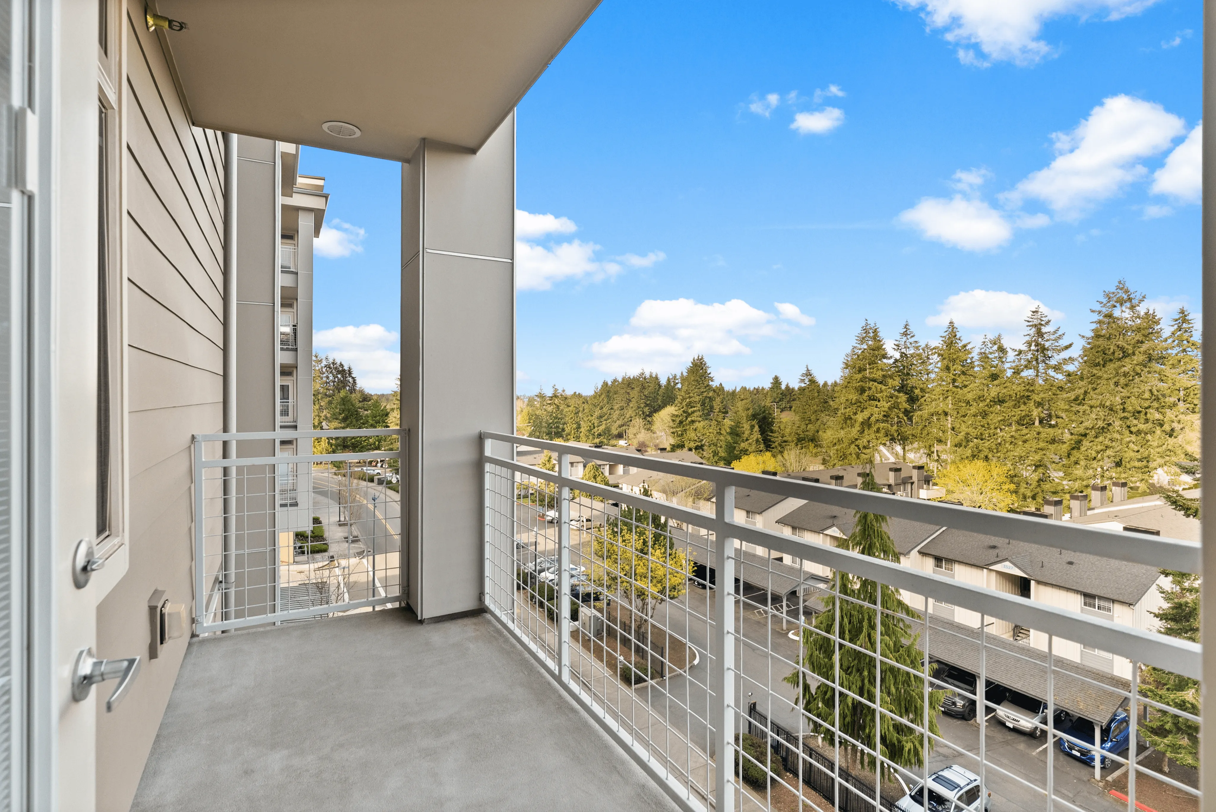 Apartment balcony with silver metal railing overlooking lush green trees under a bright blue sky with minimal clouds.