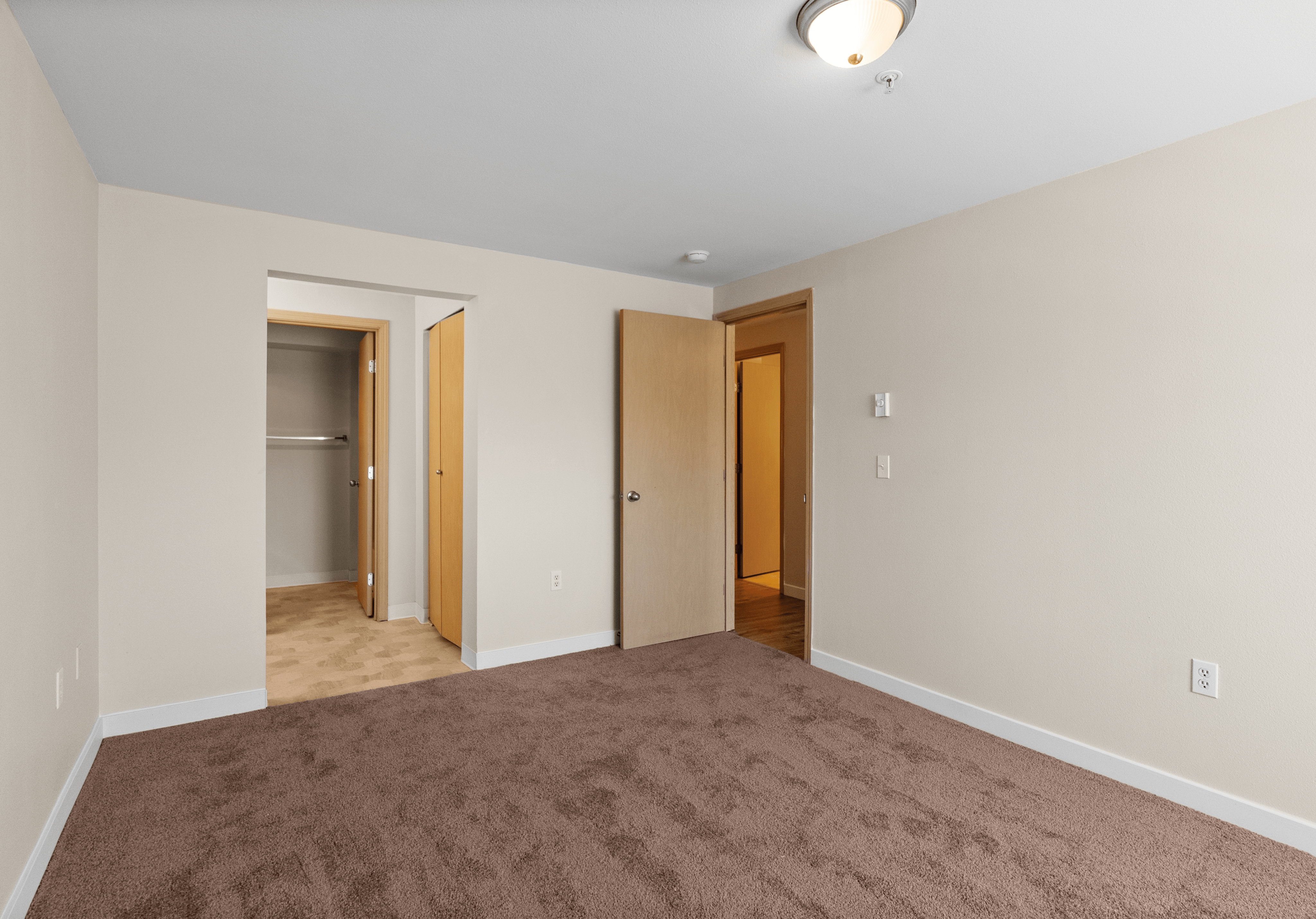 Spacious empty living room with light wood flooring, beige walls, and modern wall sconces, featuring a white door and two wooden interior doors.