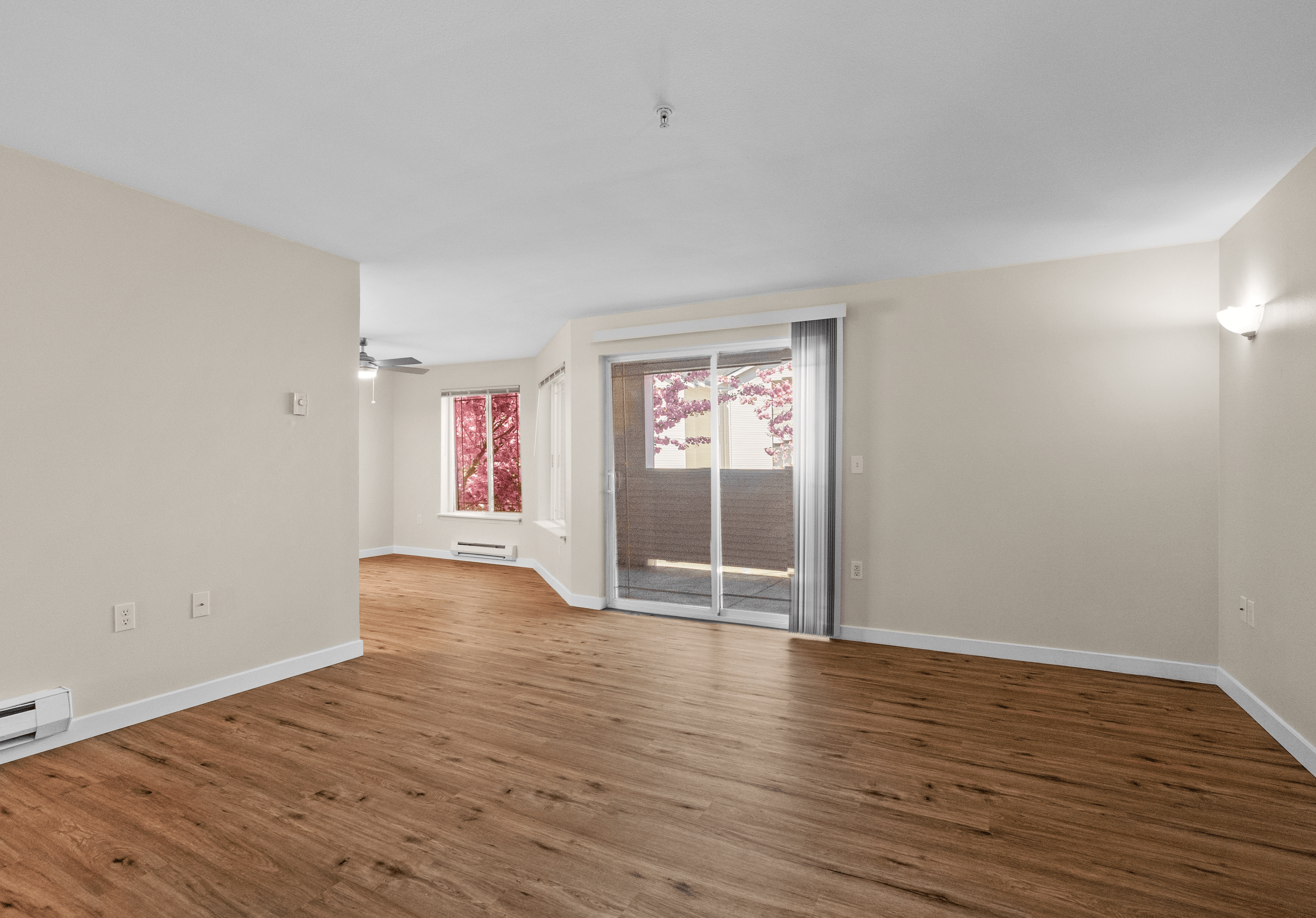 Spacious living room with light wood-style flooring, sliding glass door to a private patio, and large windows showcasing blooming pink trees