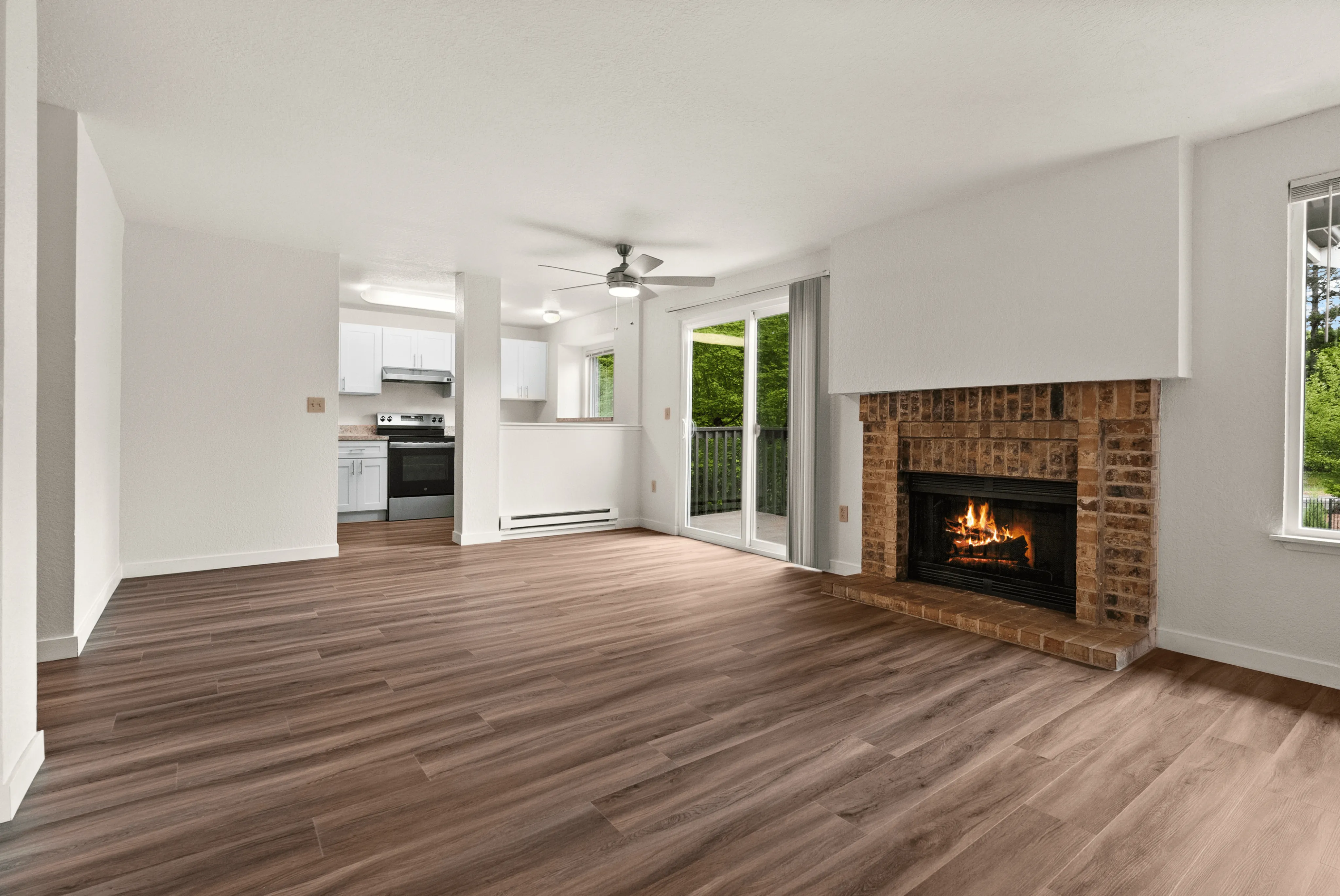 Open-concept apartment interior featuring a cozy brick fireplace, modern wood-style flooring, and a view into a bright white kitchen with stainless steel appliances.