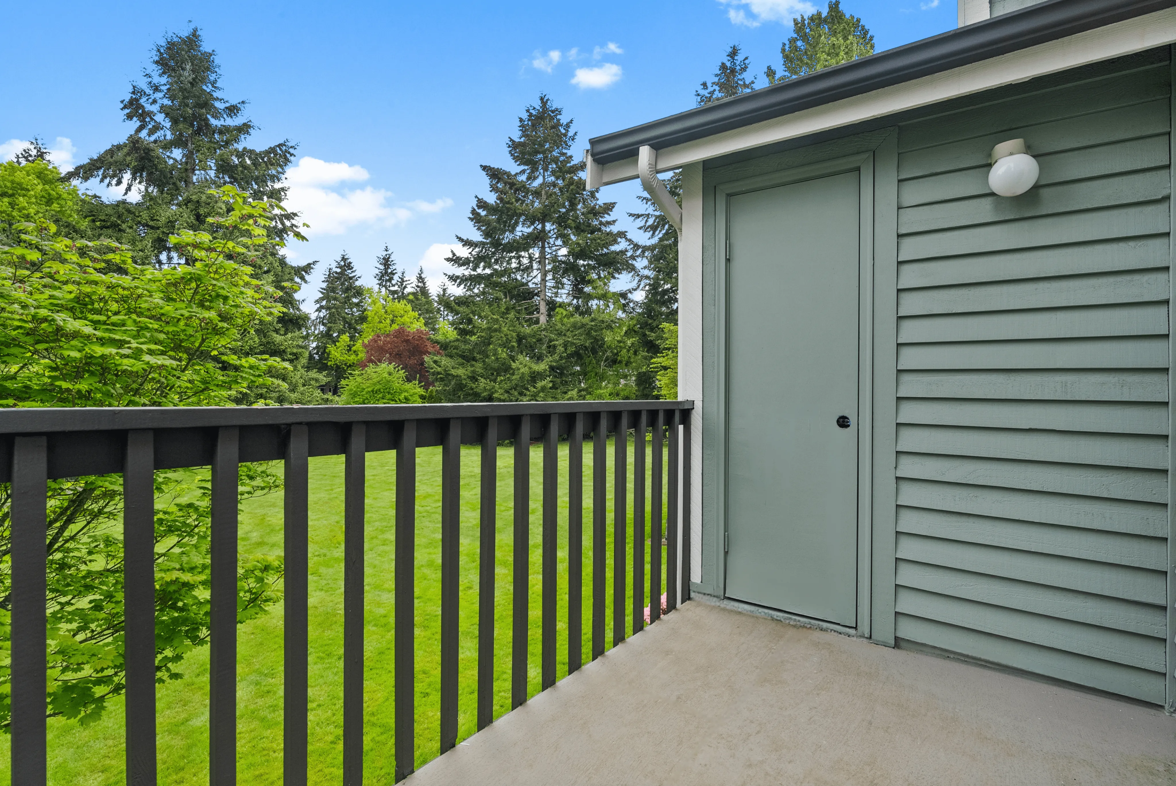 Balcony with black railing overlooking a green lawn and trees, attached to a house with green siding.