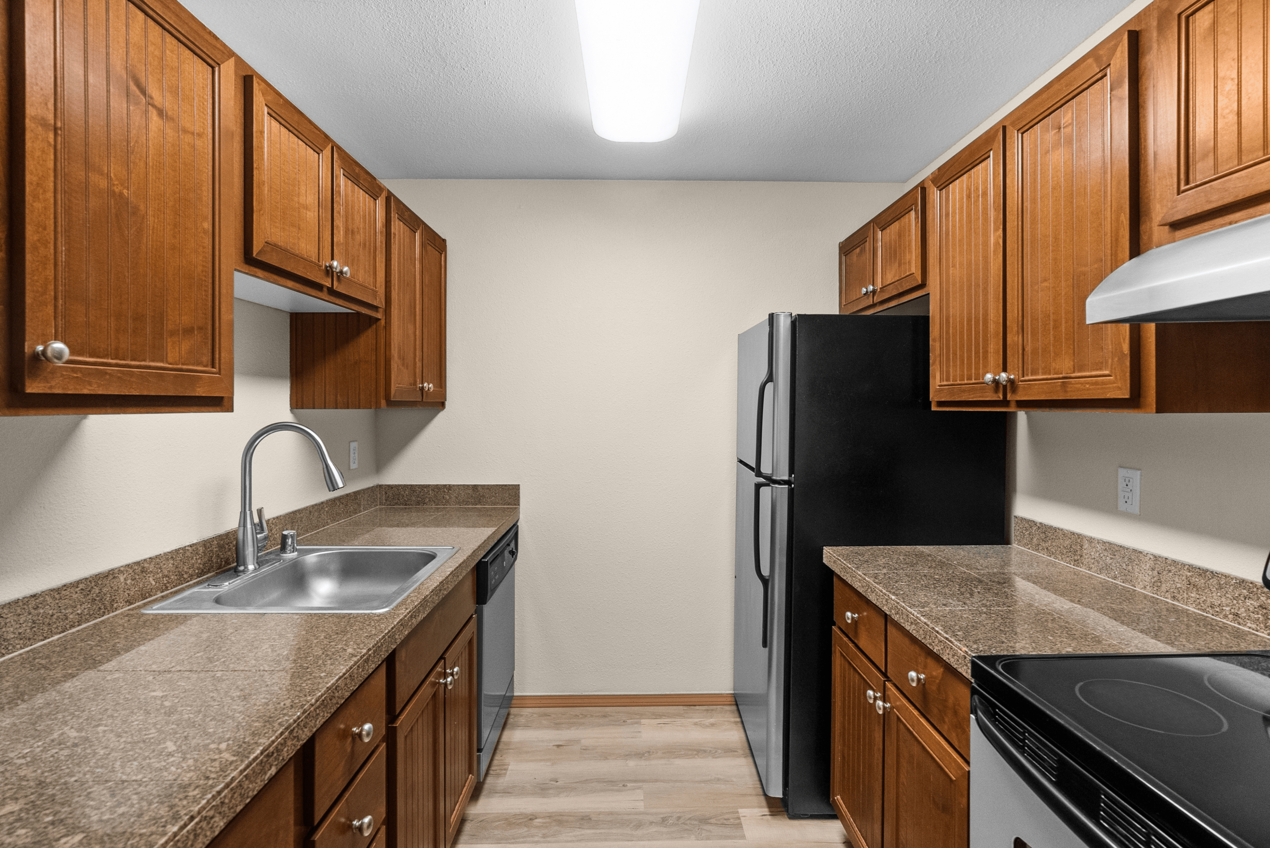 Modern galley-style kitchen with wood cabinets, granite-look countertops, stainless steel appliances, and vinyl plank flooring.