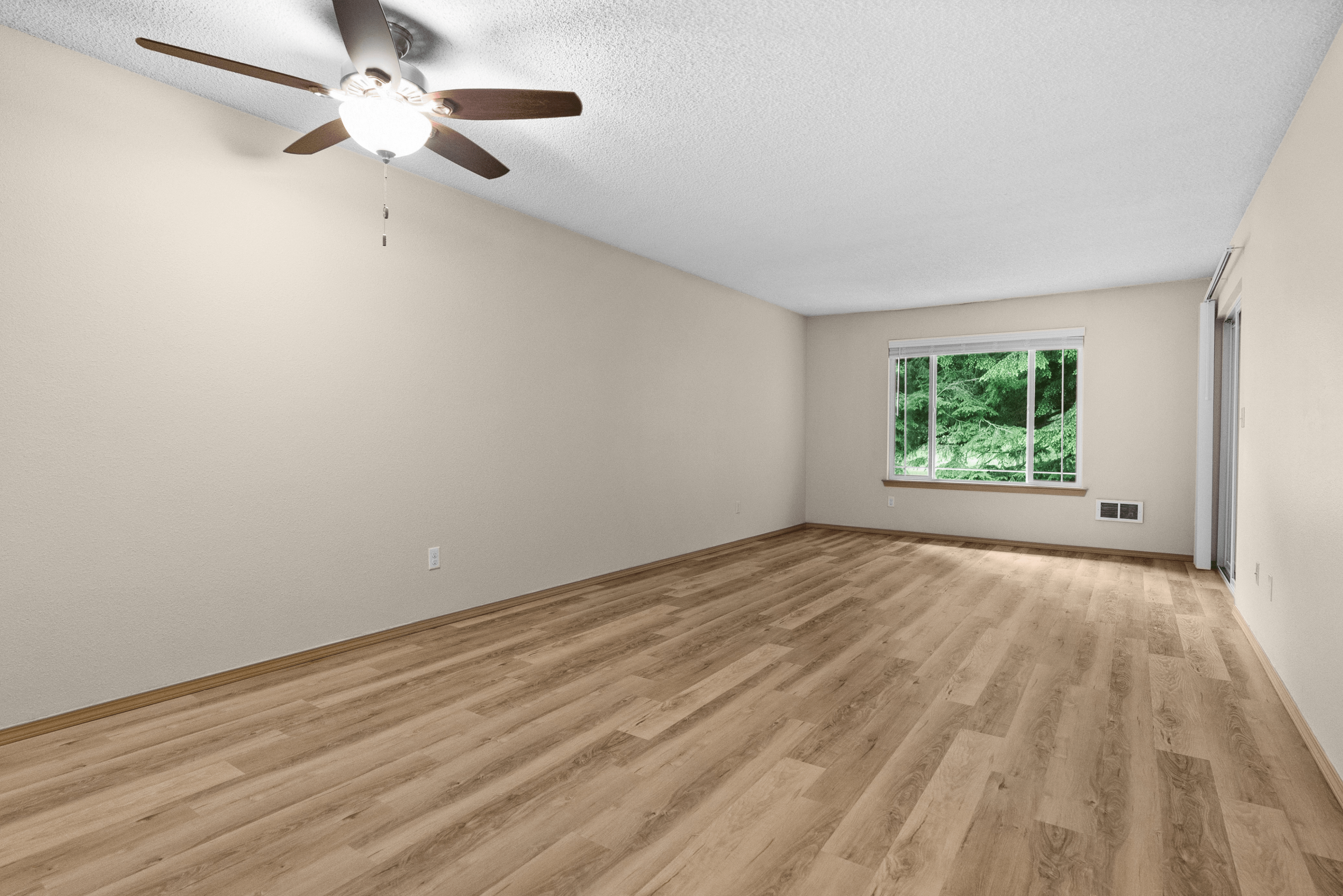 Spacious living room with wood laminate flooring, ceiling fan, and large window offering views of lush greenery outside.