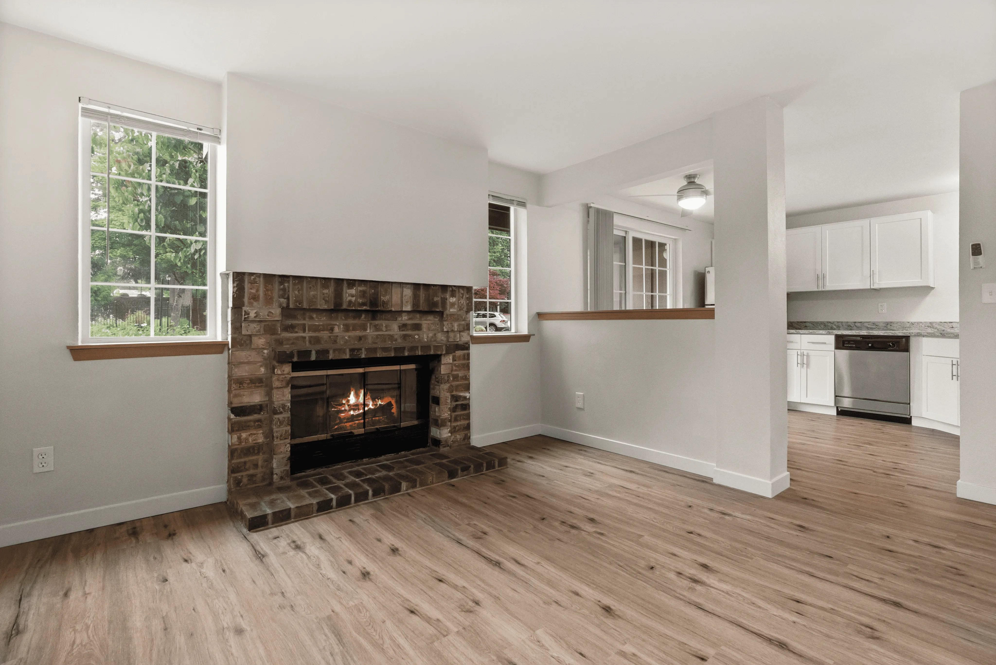 Inviting living space with a charming brick fireplace, large windows for natural light, and a glimpse into a modern kitchen with sleek cabinetry.