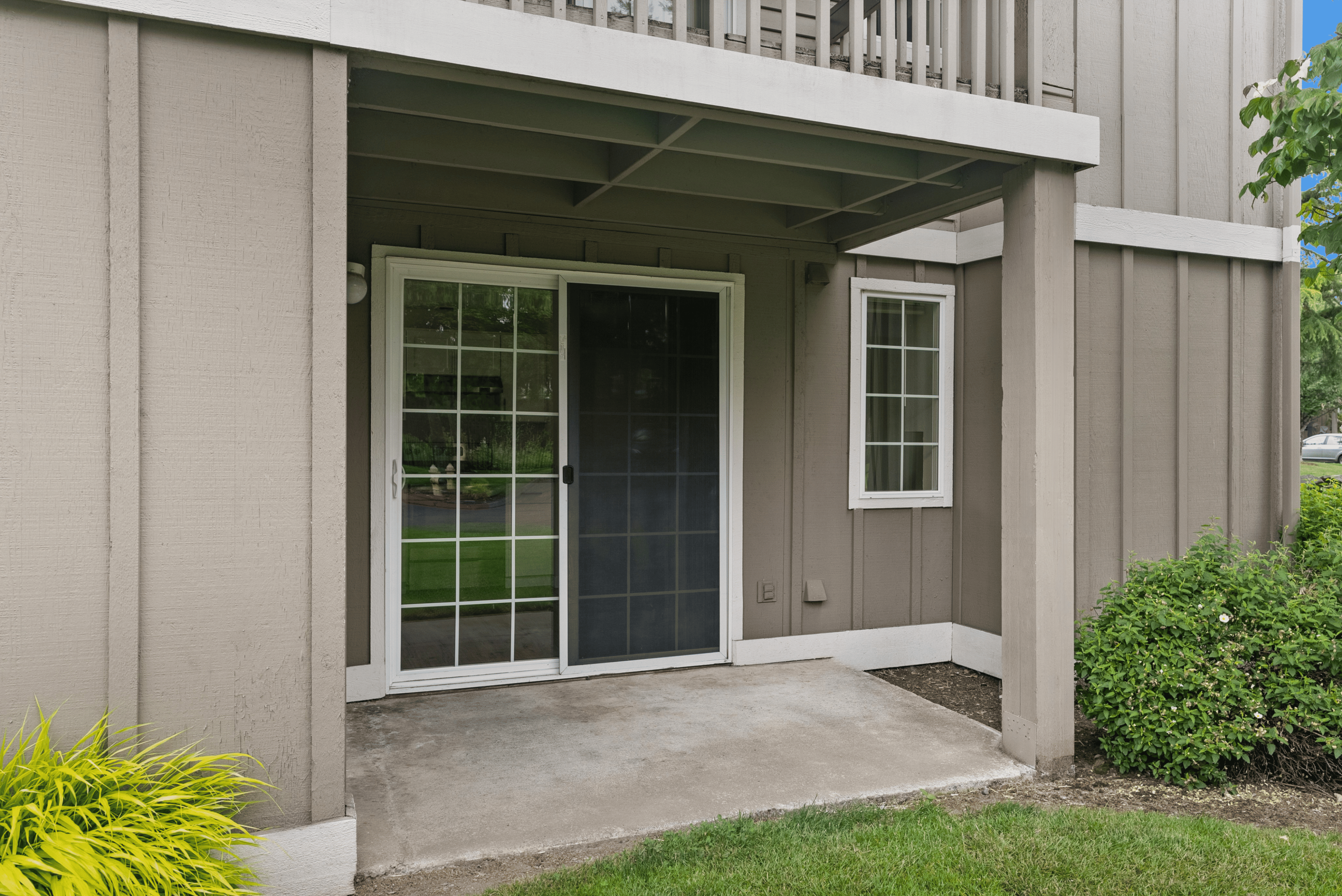 Charming outdoor patio with sleek sliding glass doors, surrounded by lush greenery and modern brown siding, perfect for relaxation and enjoying nature.