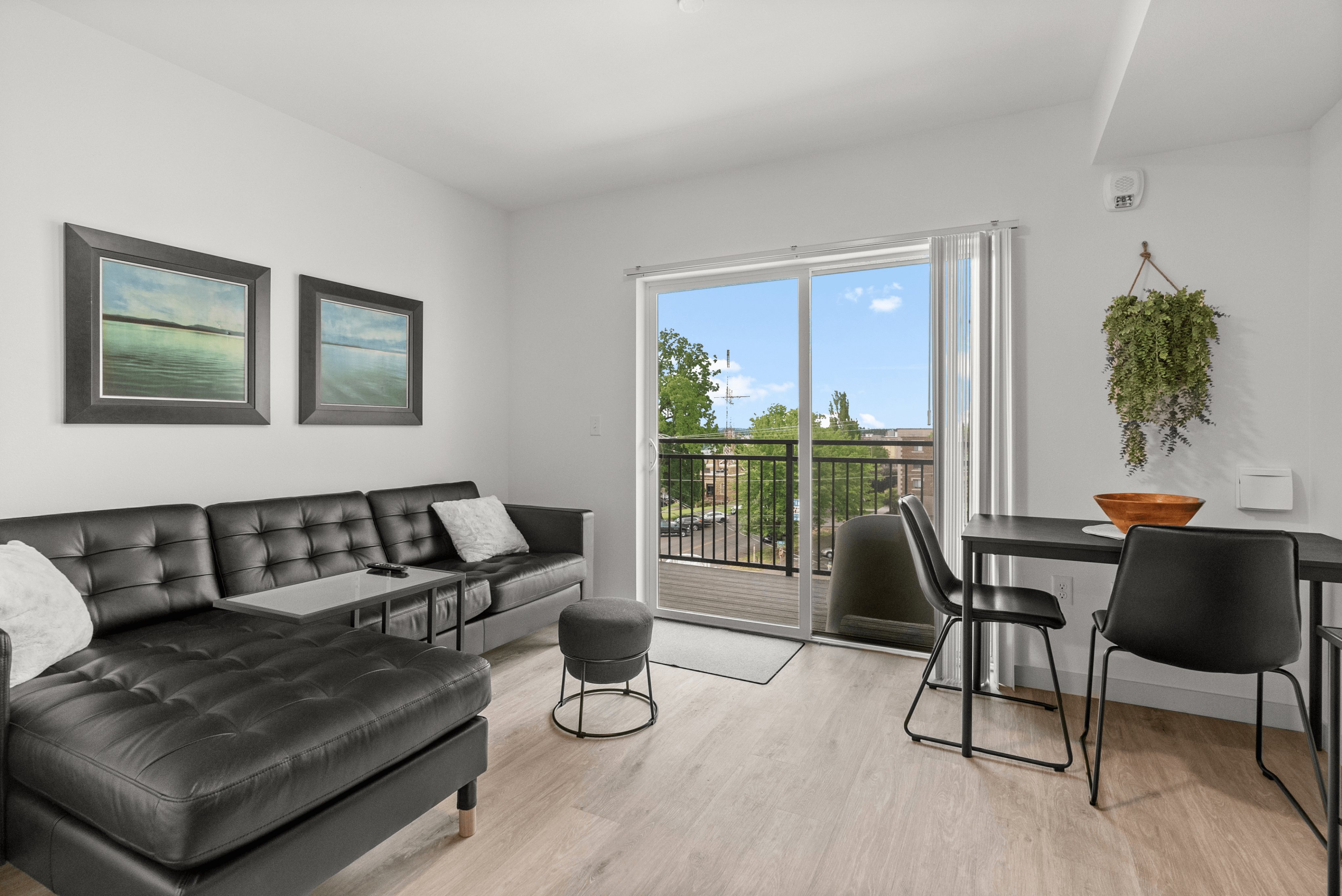 Modern living room with sleek black leather sofa, elegant artwork, and large windows opening to a balcony with a scenic view.