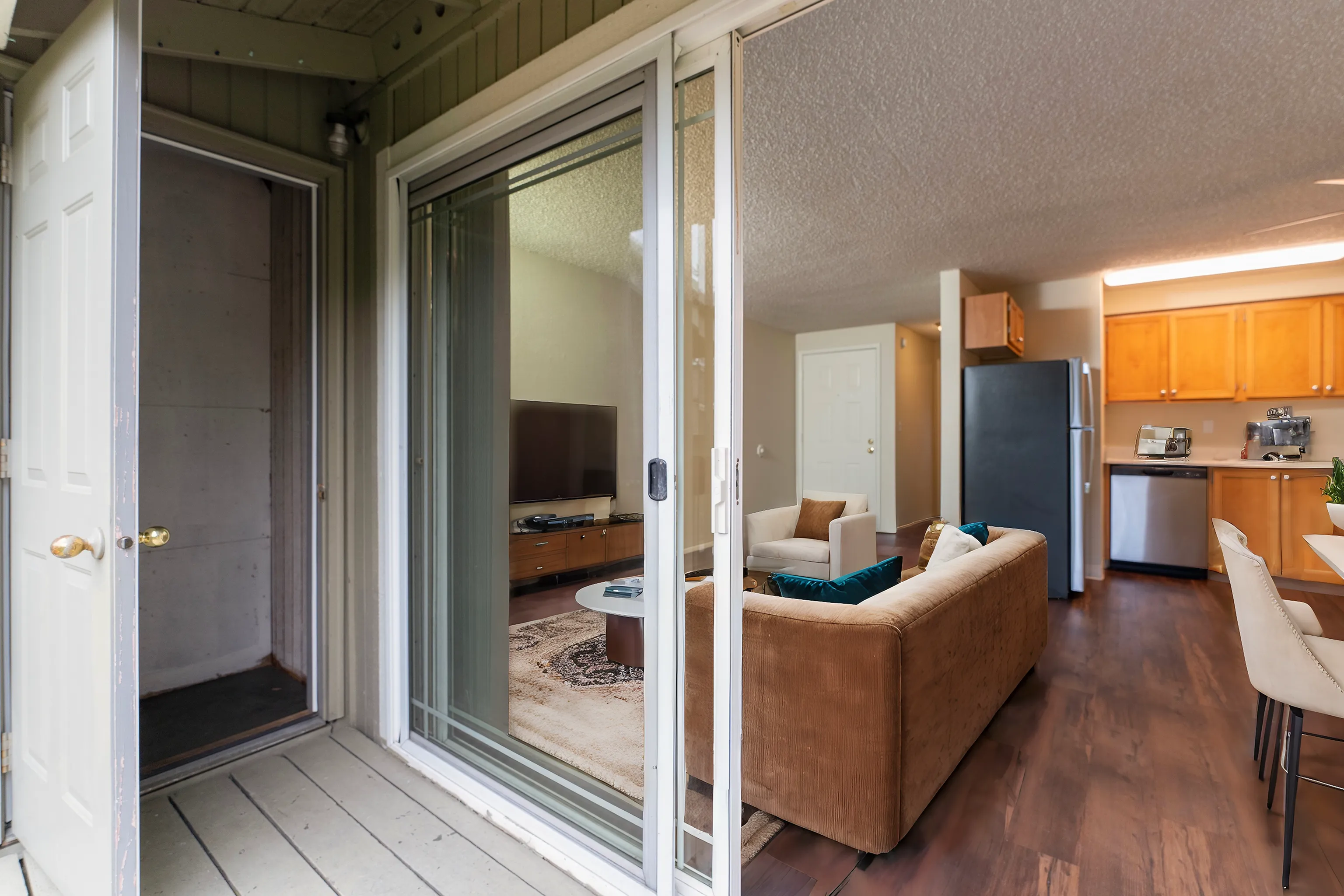 Inviting apartment with an open-concept living space, featuring a cozy living room, modern kitchen with stainless steel appliances, and sleek hardwood floors viewed through a sliding glass door.