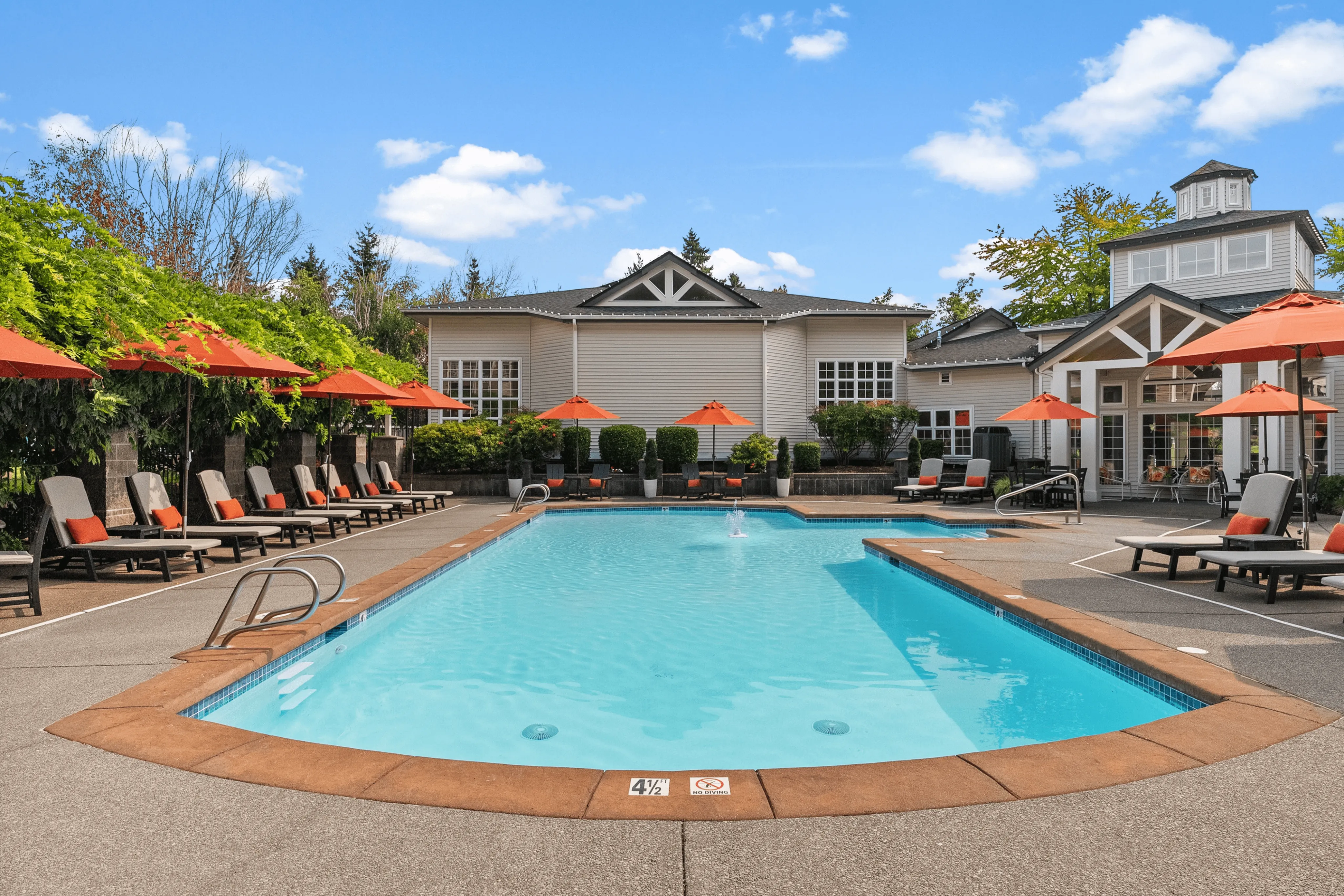 Inviting outdoor pool area with elegant lounge chairs and vibrant orange umbrellas, set against a modern clubhouse with lush greenery, perfect for relaxation.