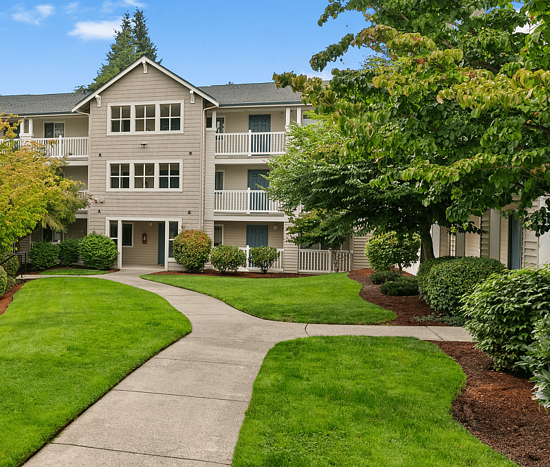 Charming apartment complex with a welcoming entrance, lush green landscaping, and bright balconies set against a clear blue sky.