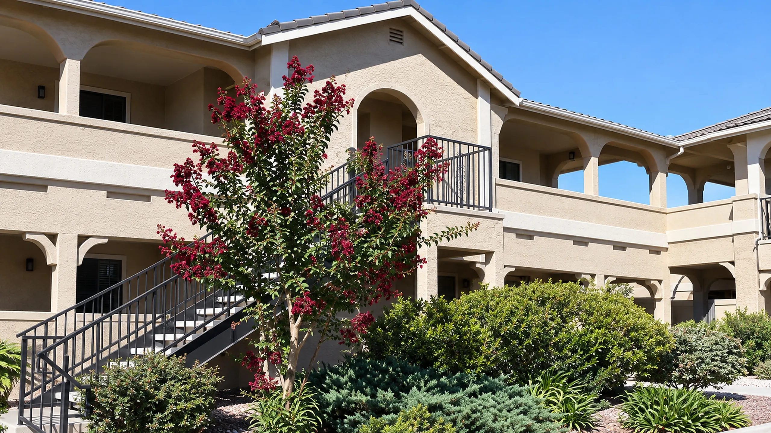Charming two-story apartment building with arched balconies, surrounded by vibrant landscaping and clear blue skies.