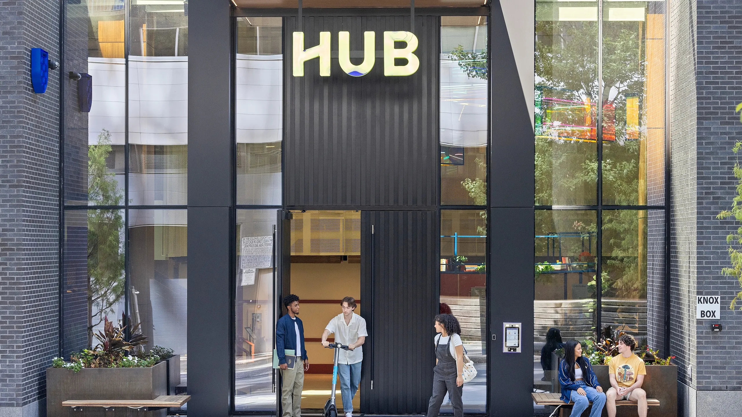 Modern apartment entrance with sleek design under HUB sign, featuring engaged pedestrians and vibrant, urban environment.