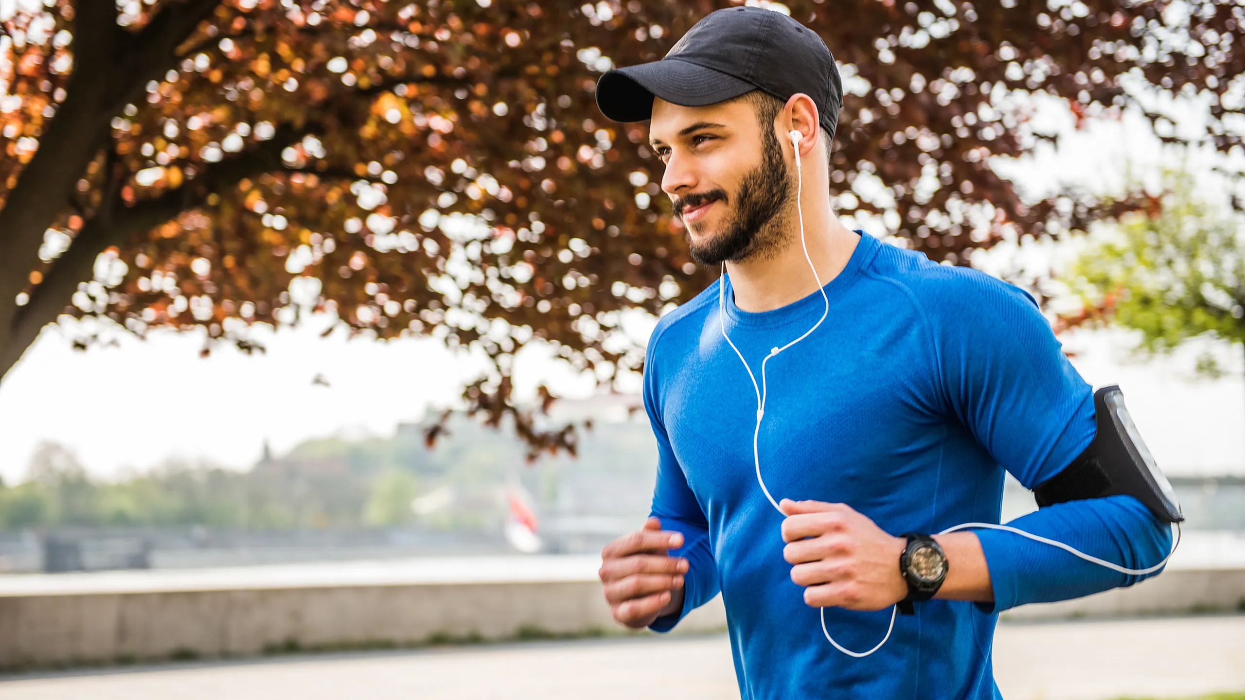 Man in blue athletic wear jogging with earphones, enjoying a run in a scenic park with autumn foliage.