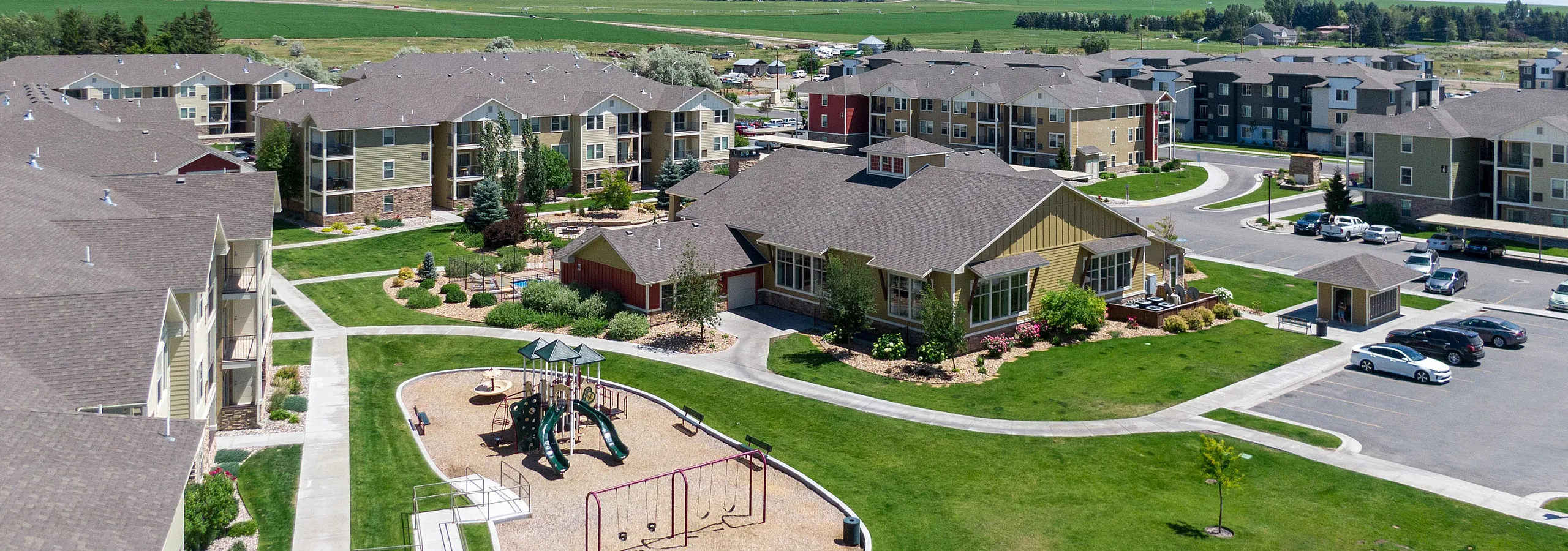 Charming aerial view of a modern apartment complex with lush green spaces, playground, and ample parking. A picturesque community setting surrounded by scenic landscapes.