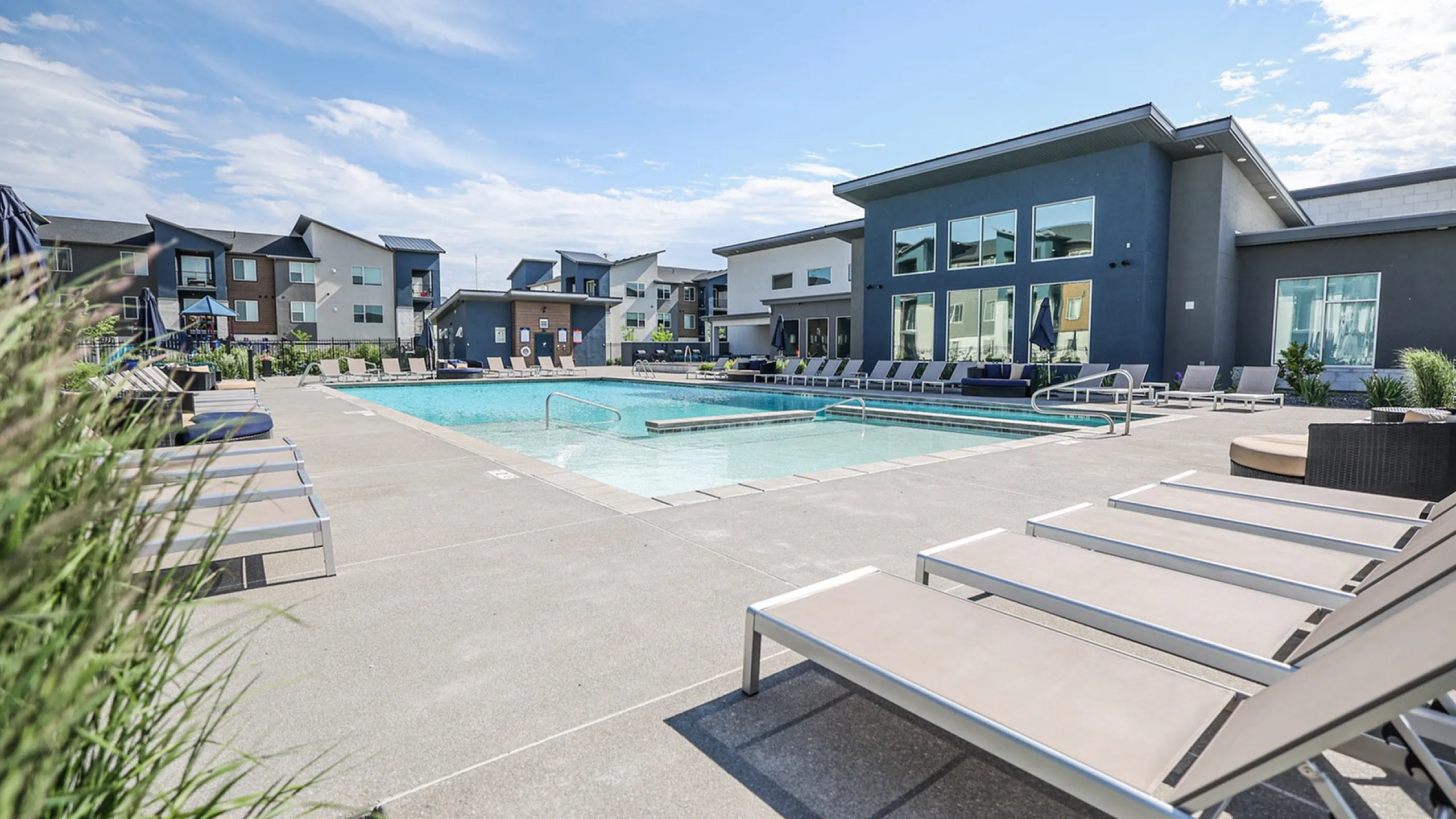 Elegant apartment complex with a luxurious outdoor pool area, surrounded by modern lounge chairs and scenic landscaping under a clear blue sky.