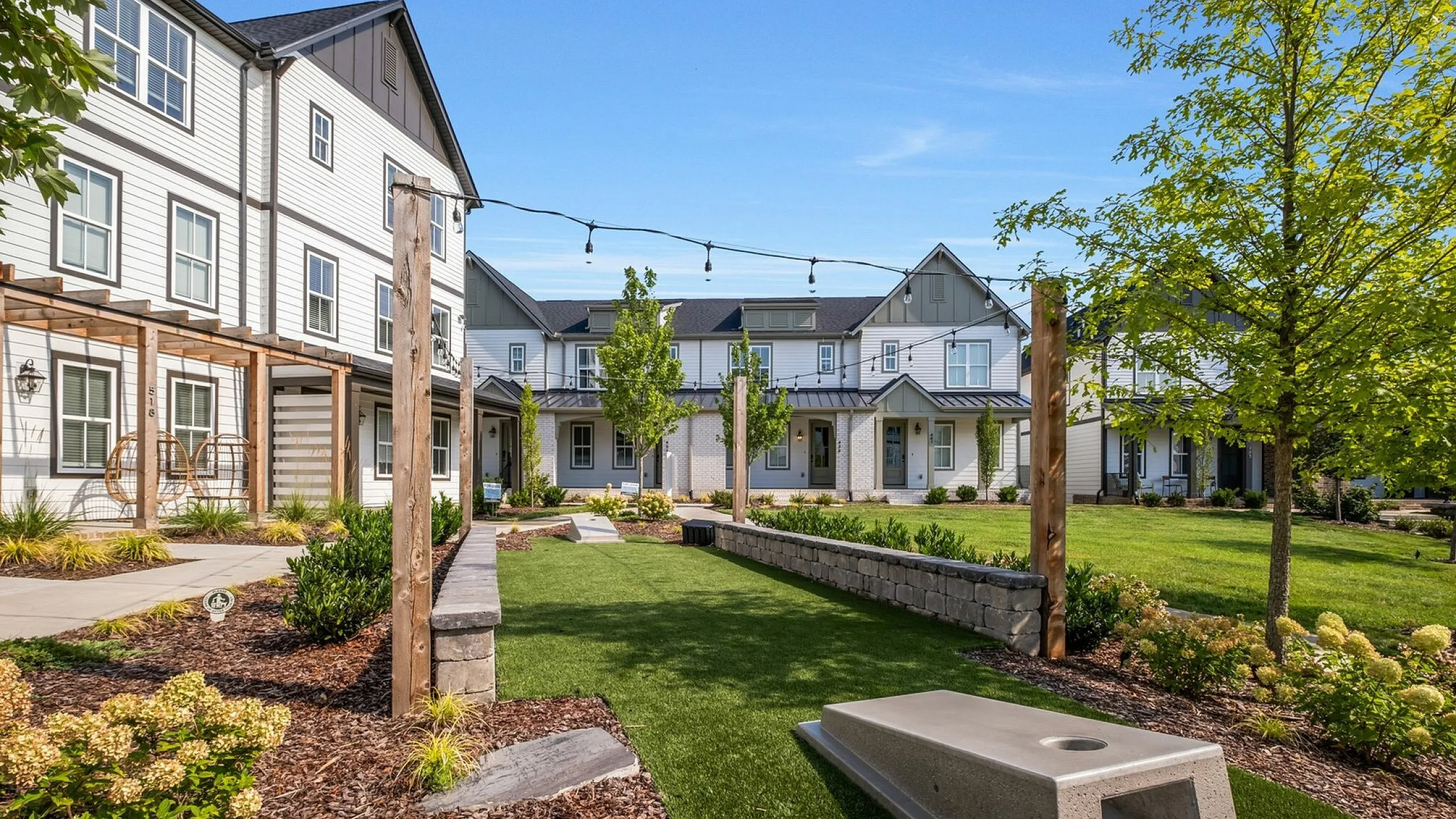 Charming outdoor space in a modern townhouse community featuring a lush green lawn, garden beds, and hanging chairs under a pergola, perfect for relaxing and socializing.